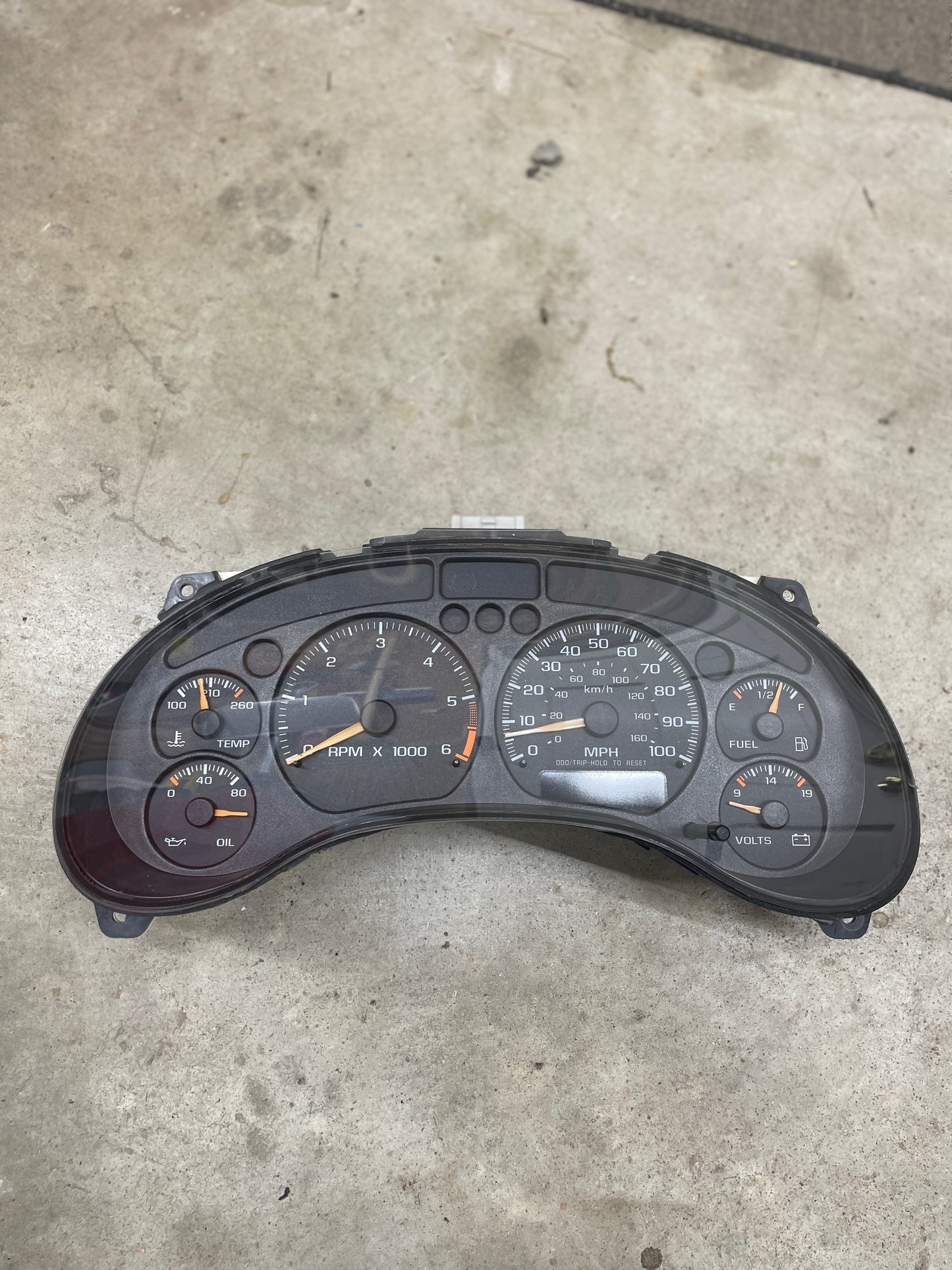 A General Motors OEM Tested Instrument Cluster with Tachometer for 2001-2005 Chevy S10 or Blazer with Console Shifter rests on concrete, its gauges—RPM, speedometer, fuel, temp, oil, voltage—clearly visible with needles at idle.