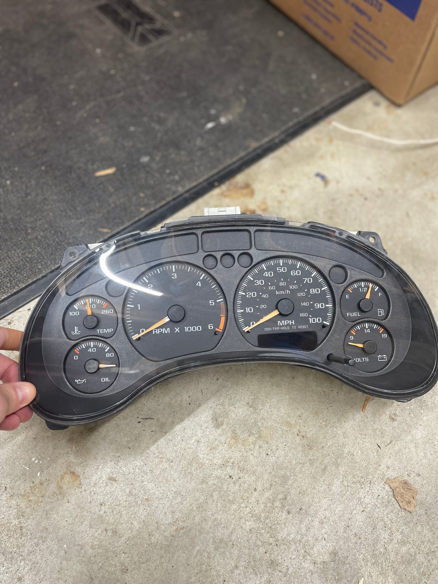 A hand holds a General Motors OEM Tested Instrument Cluster with Tachometer for 2001-2005 Chevy S10 or Blazer with Console Shifter, showing speedometer, fuel, temp, and oil pressure gauges, resting on an indoor concrete floor.