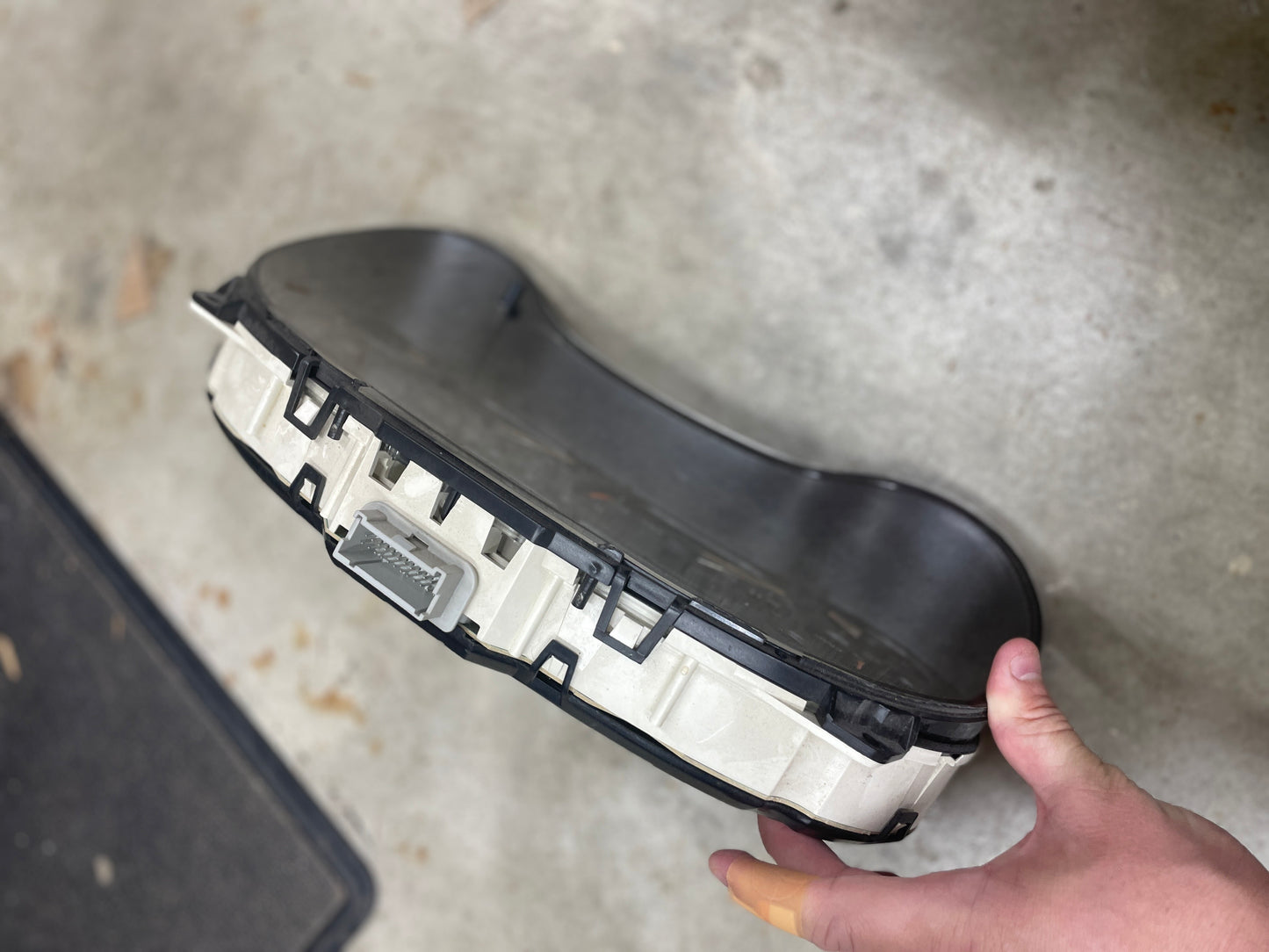 A hand holds a General Motors OEM Tested Instrument Cluster with Tachometer for 2001-2005 Chevy S10 or Blazer with Console Shifter, showing its connector plug, above a concrete floor scattered with debris.