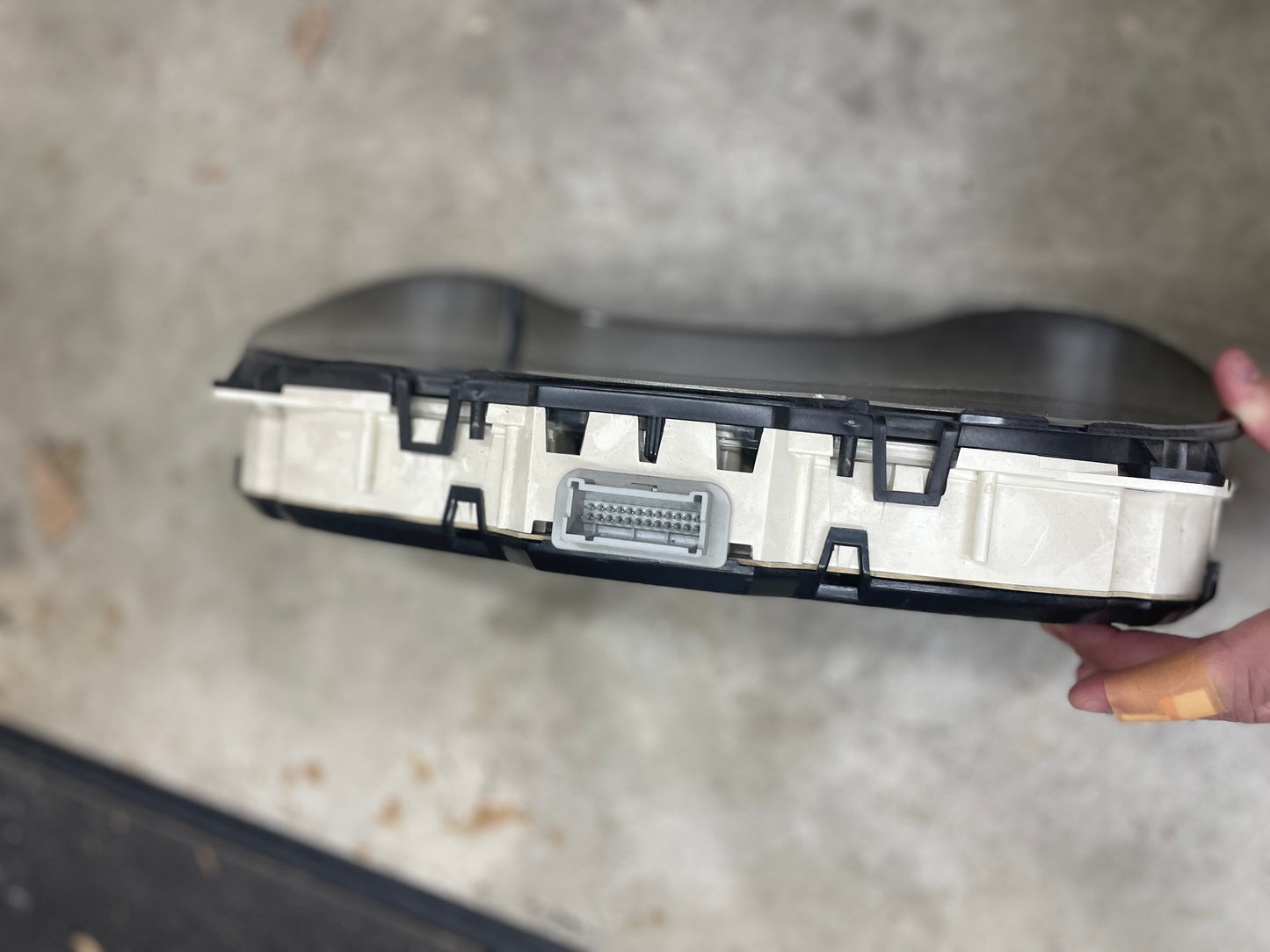 A hand holds a General Motors OEM Tested Instrument Cluster with Tachometer for 2001-2005 Chevy S10 or Blazer (console shifter), showing its multi-pin connector against a concrete background.