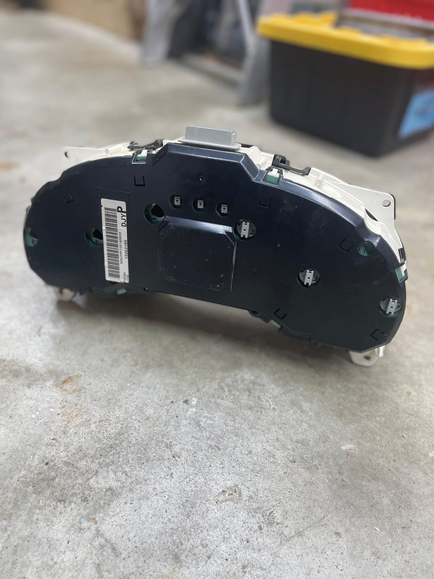 A General Motors OEM Tested Instrument Cluster with Tachometer for 2001-2005 Chevy S10 or Blazer with Console Shifter is placed face down on a concrete floor, with a yellow and black storage bin in the background.