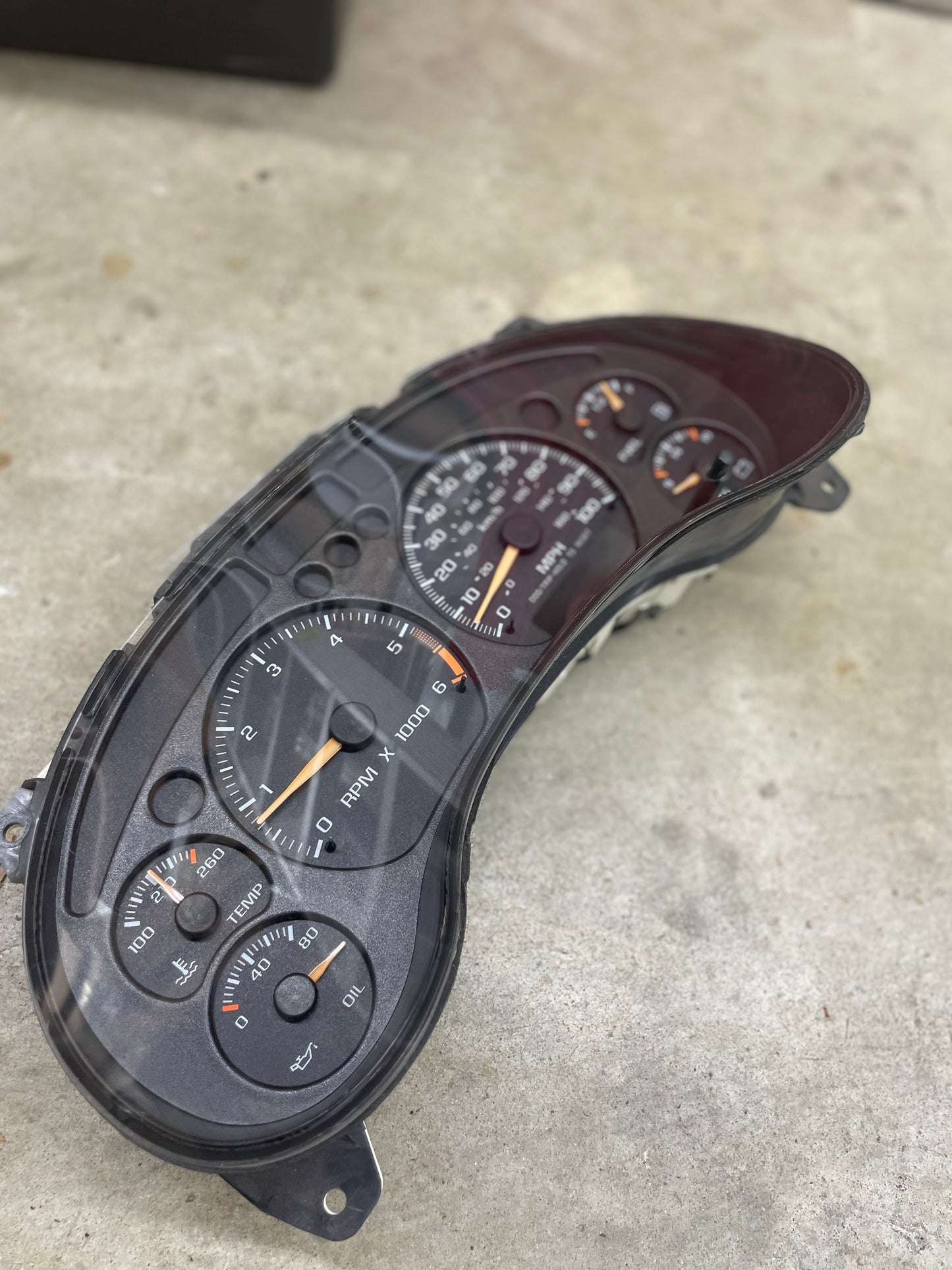 A close-up of a General Motors OEM Tested Instrument Cluster with Tachometer, designed for 2001-2005 Chevy S10 or Blazer with console shifter, featuring speedometer, fuel and oil gauges, shown on a concrete floor.