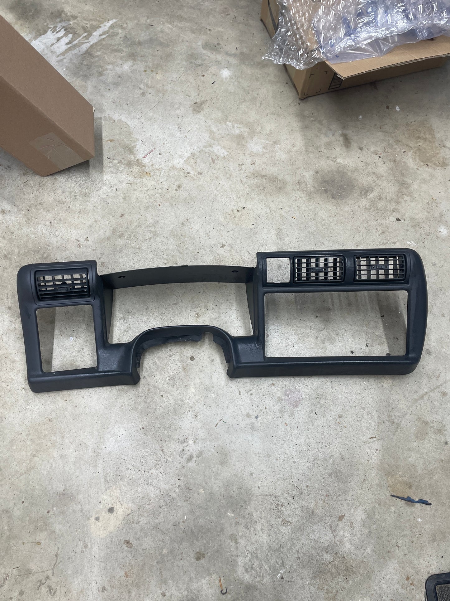 A General Motors OEM Instrument Cluster Dash Bezel in dark gray for 1995-1997 Chevy S-10, Blazer, and more, with air vents, rests on a concrete floor surrounded by cardboard boxes and packing material.