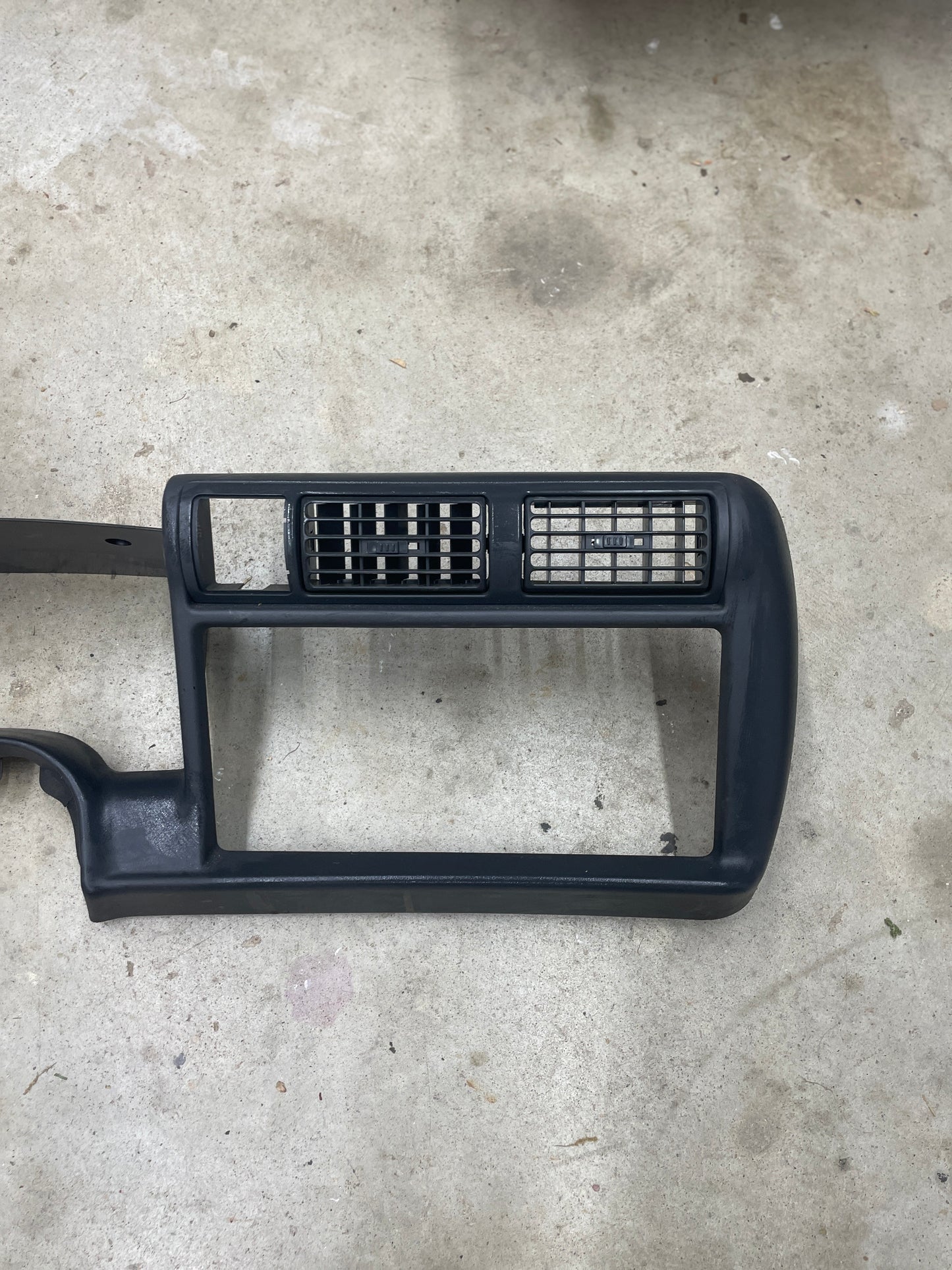 A General Motors OEM dark gray instrument cluster dash bezel for 1995-1997 Chevy S-10, Blazer, and more, featuring two rectangular vents and a lower open space, is shown on a light concrete floor.