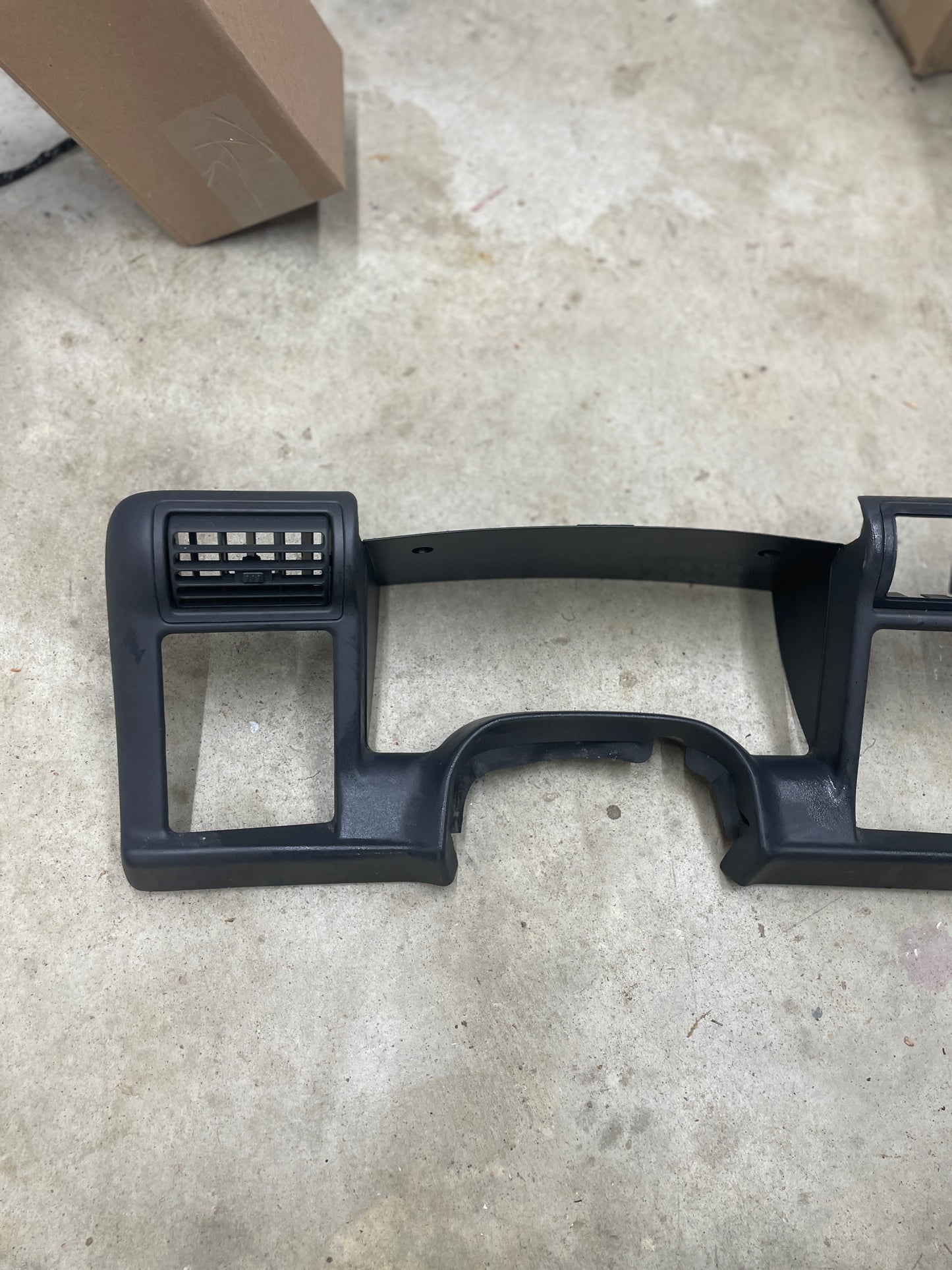 A General Motors OEM Instrument Cluster Dash Bezel in dark gray for 1995-1997 Chevy S-10, Blazer, and similar models lies on a concrete floor. A rectangular air vent is on the left; a cardboard box is partially seen in the top left.