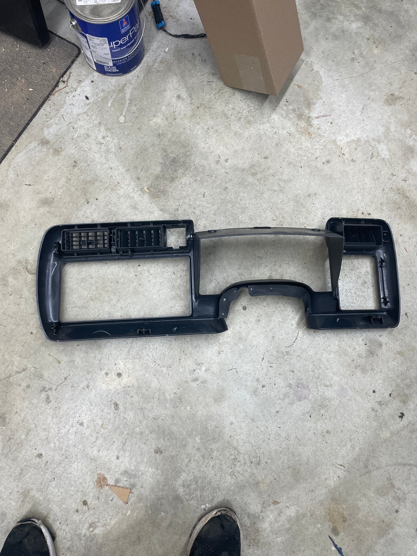A General Motors OEM dark gray instrument cluster dash bezel for 1995-1997 Chevy S-10, Blazer, and more rests on a concrete floor. Paint can, cardboard box, and shoes are seen at the edges.