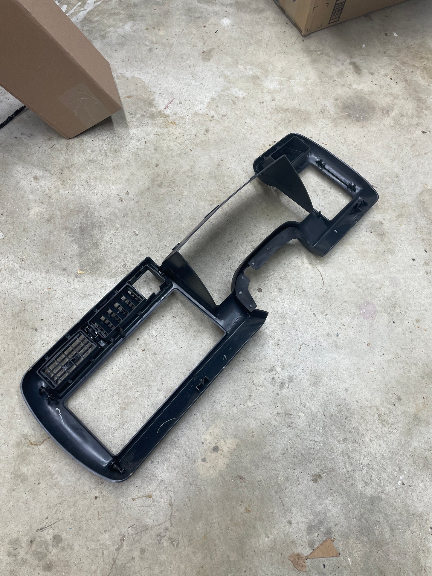 A General Motors OEM Instrument Cluster Dash Bezel in dark gray, designed for 1995-1997 Chevy S-10, Blazer and more, has vent and control cutouts and is shown lying on a concrete floor with cardboard boxes in the background.