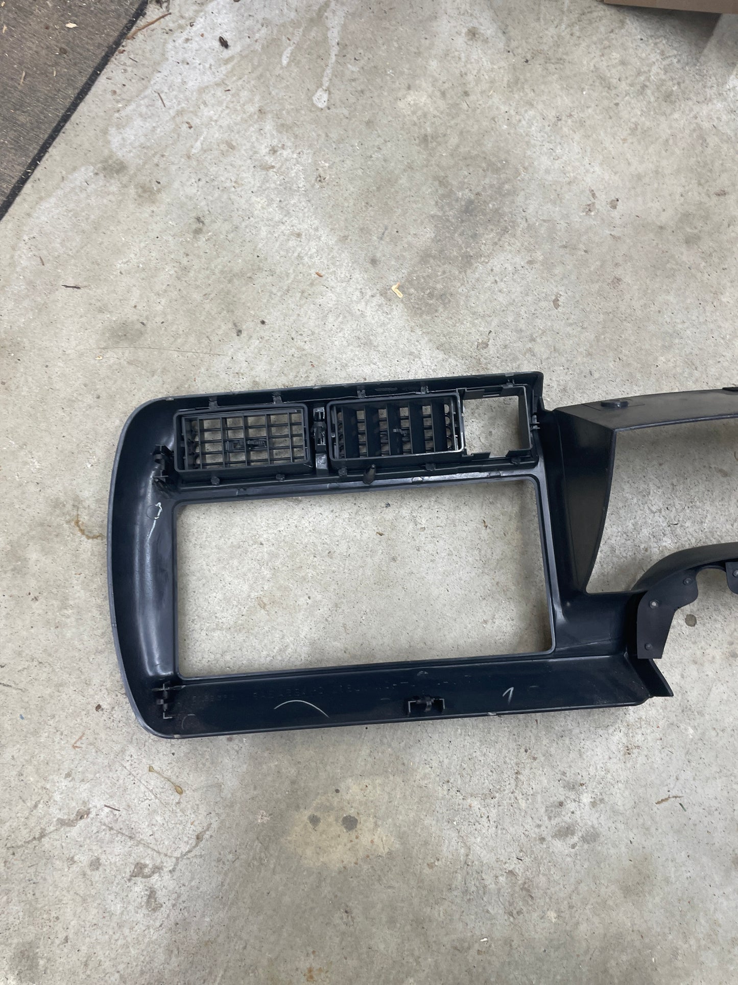 A General Motors OEM instrument cluster dash bezel in dark gray for 1995-1997 Chevy S-10 and Blazer is lying vent side up on a concrete floor. The plastic trim appears slightly worn.
