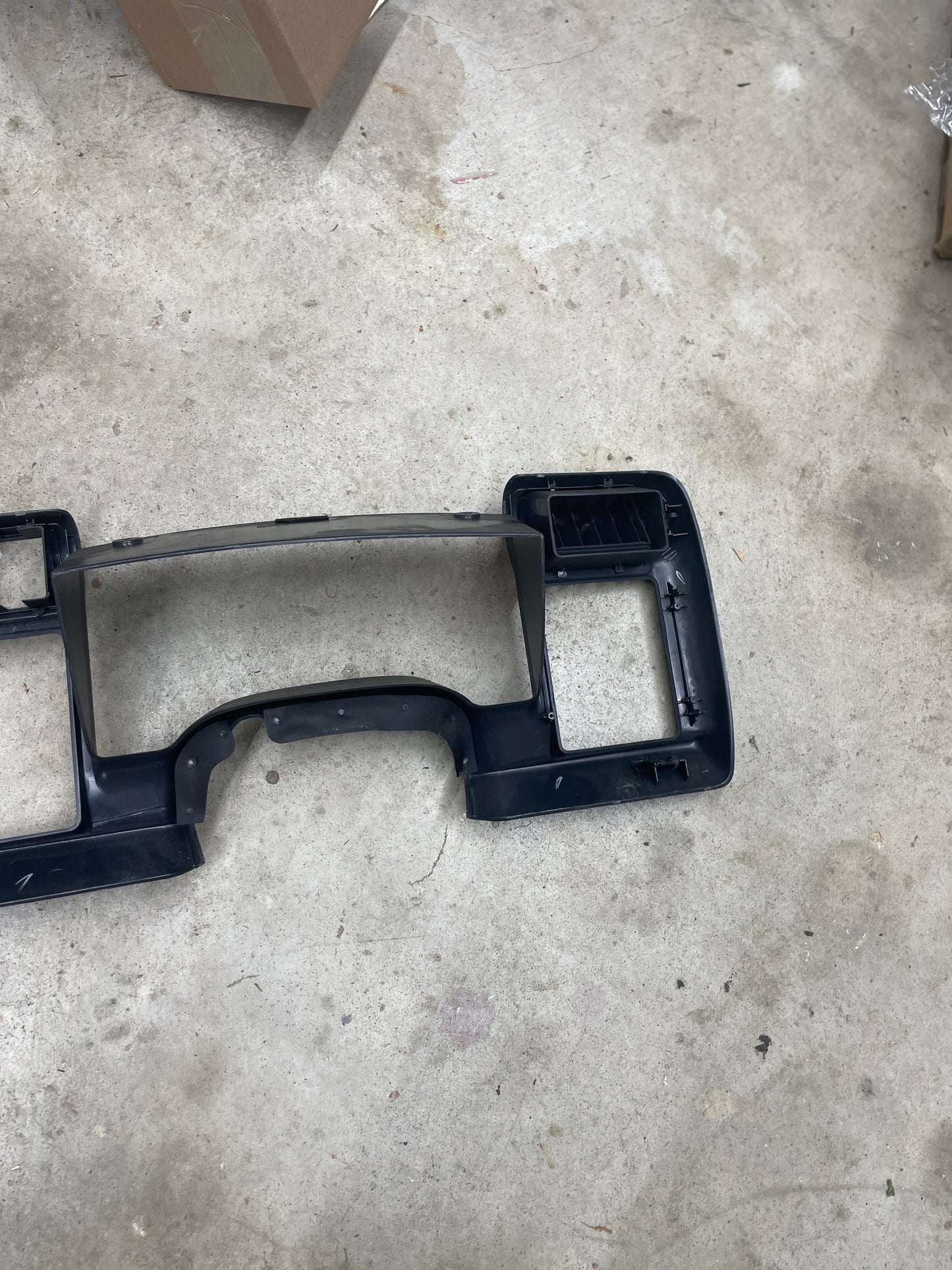 A General Motors OEM Instrument Cluster Dash Bezel in dark gray for 1995-1997 Chevy S-10, Blazer, and more rests on a workshops concrete floor, showing some marks with cardboard boxes nearby.