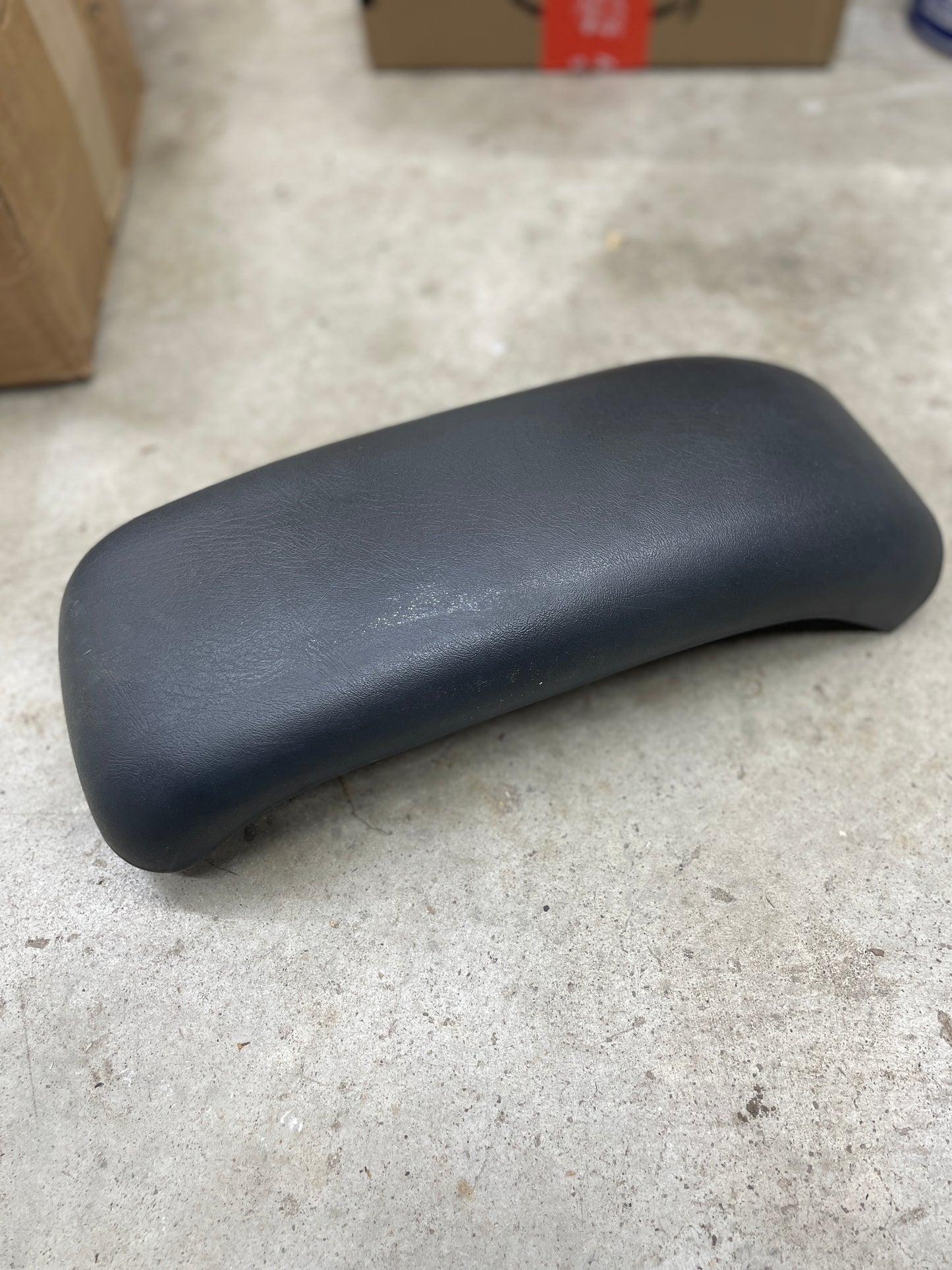 A General Motors OEM Center Lid Armrest in dark gray for Auto Trans Shifter Console (1996-2005 Chevy S10, Blazer, and more) rests on concrete flooring with cardboard boxes visible in the background.