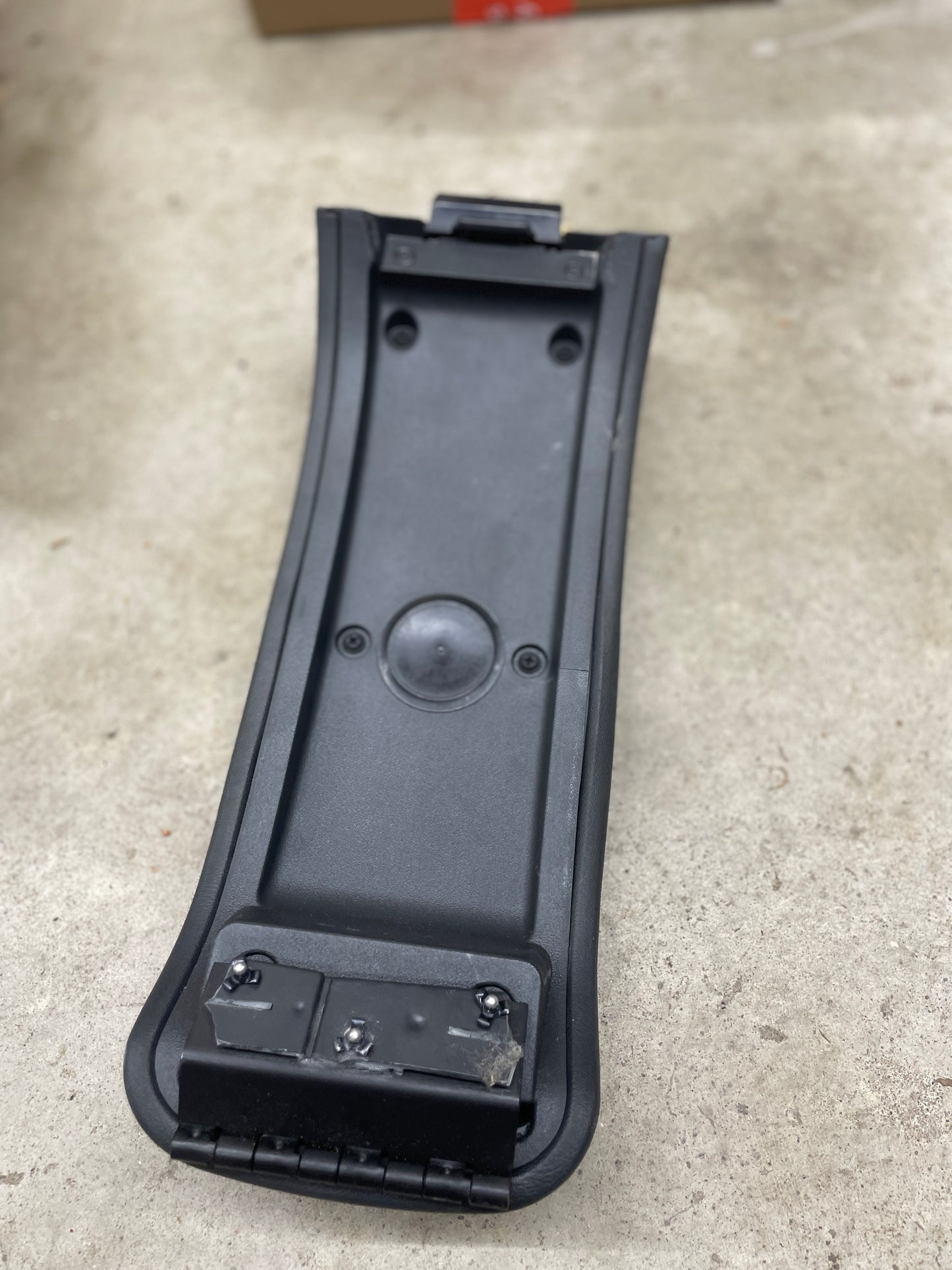 A General Motors OEM Center Lid Armrest in dark gray for 1996-2005 Chevy S10 and Blazer automatic shifter consoles rests on a concrete floor, showing its hinges, mounting points, raised circular section, and rectangular attachment points.