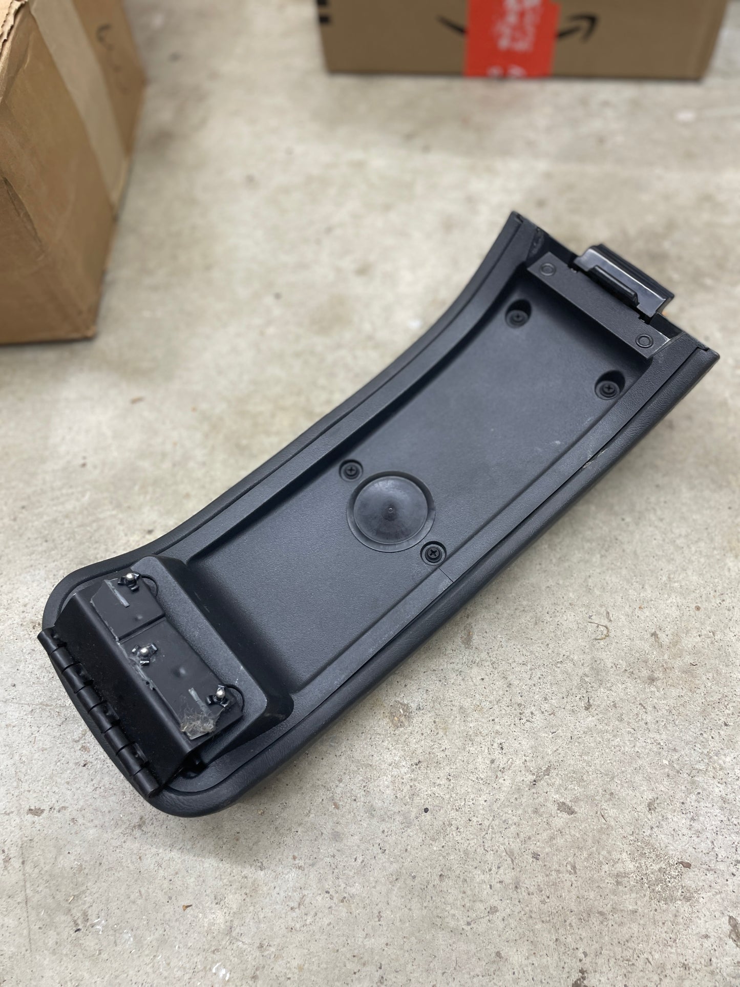 A General Motors OEM Center Lid Armrest in dark gray for auto trans shifter console (1996-2005 Chevy S10, Blazer, and more) with screws and attachment points lies on a concrete floor near cardboard boxes.