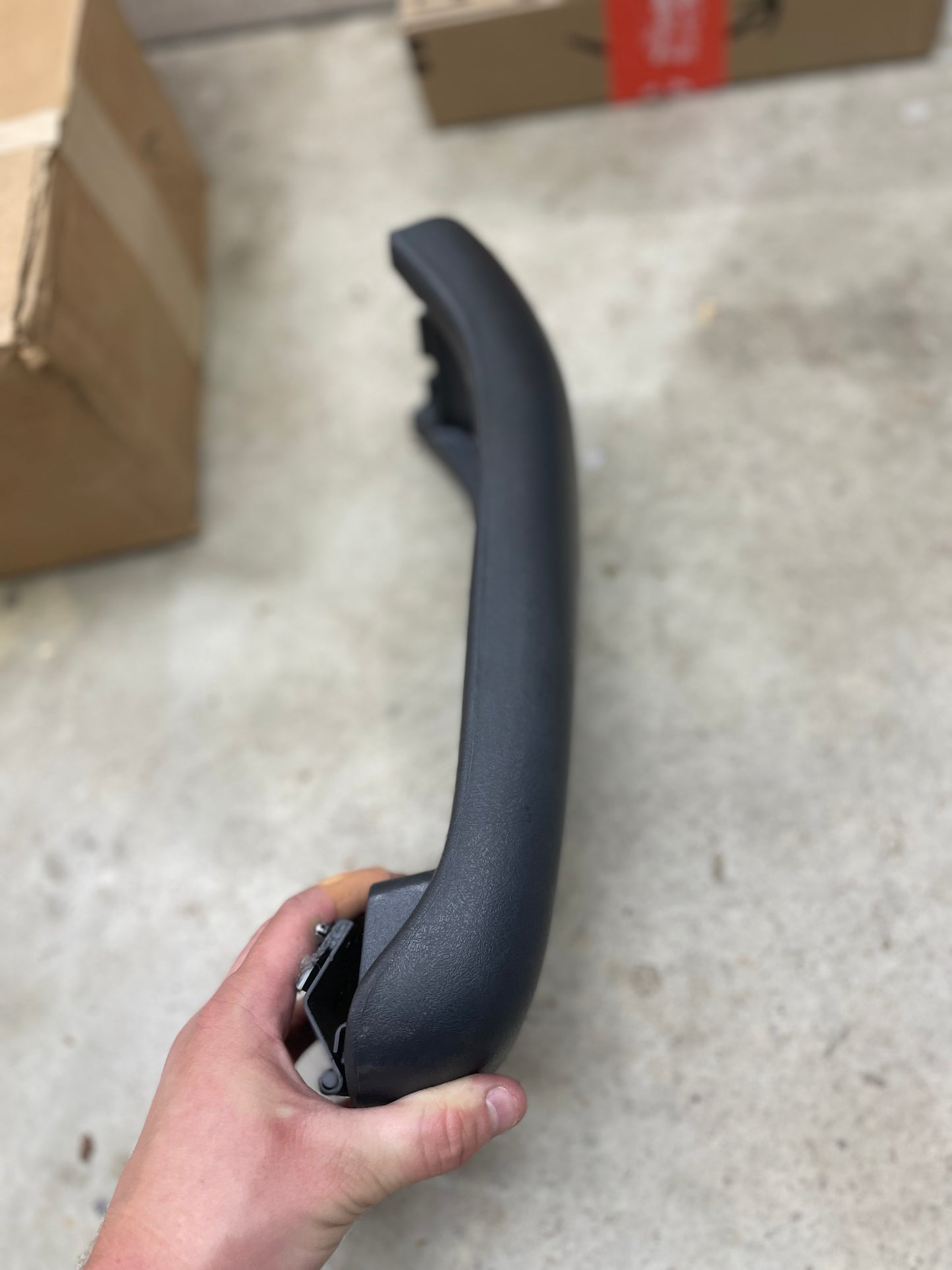 A hand holds a dark gray General Motors OEM Center Lid Armrest for 1996-2005 Chevy S10 Auto Trans Console, over concrete with boxes in the background—ideal for replacing or upgrading your factory center console.