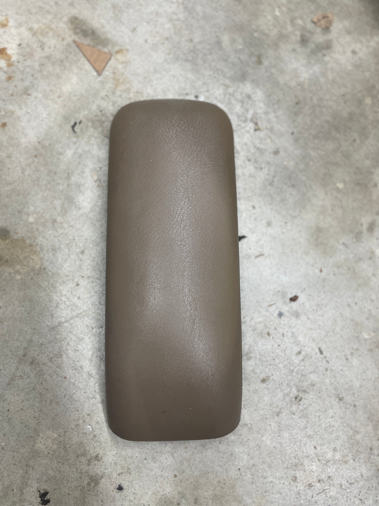 A General Motors OEM Center Lid Armrest in tan for auto trans shifter console (1996-2005 Chevy S10, Blazer, etc.) rests on a rough light gray concrete surface with scattered debris and stains nearby.