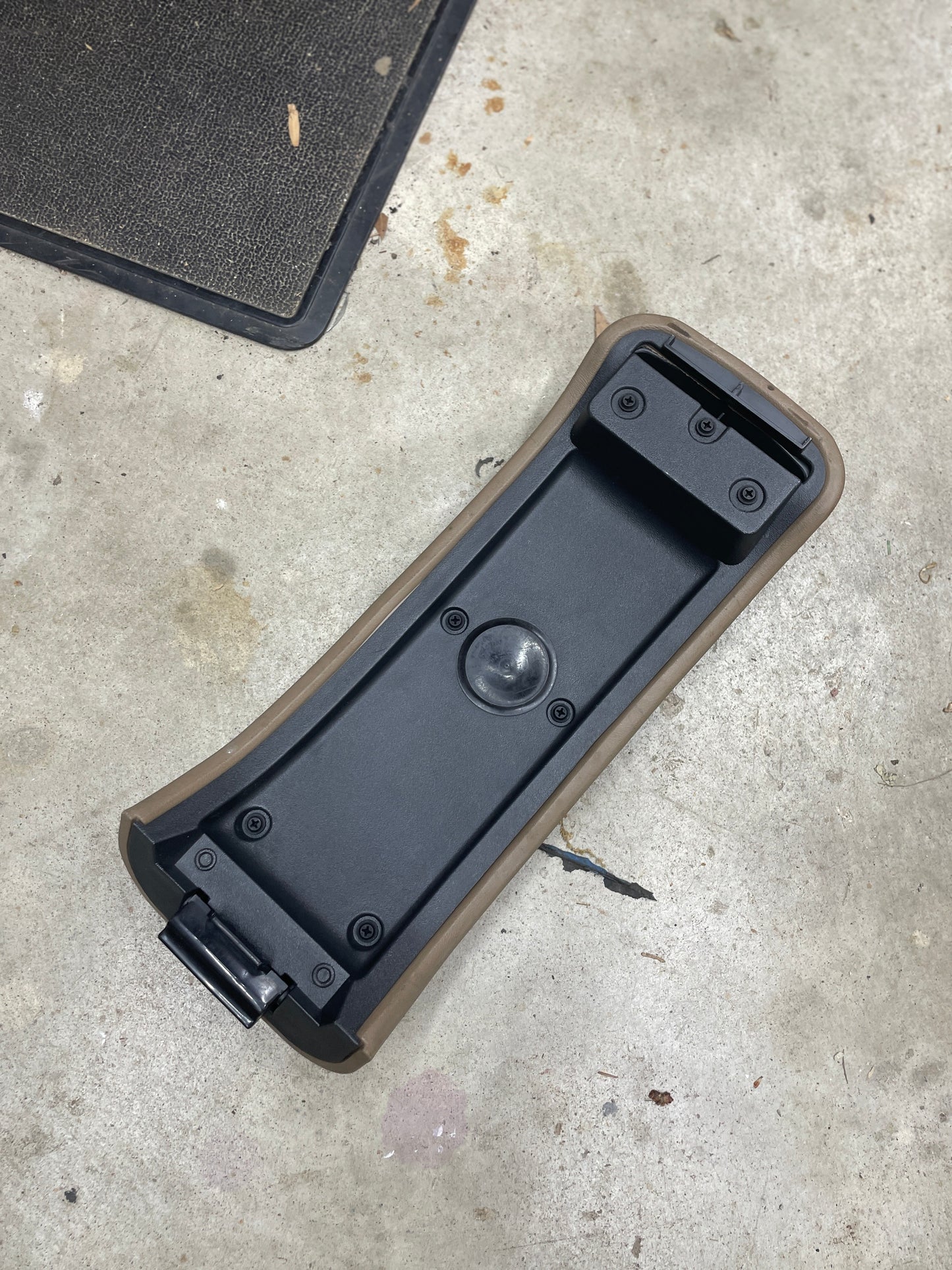 A tan plastic OEM Center Lid Armrest by General Motors for auto trans shifter consoles (1996-2005 Chevy S10, Blazer, and more) lies face up on a concrete floor, with part of a black mat visible. Ideal for replacing damaged originals.