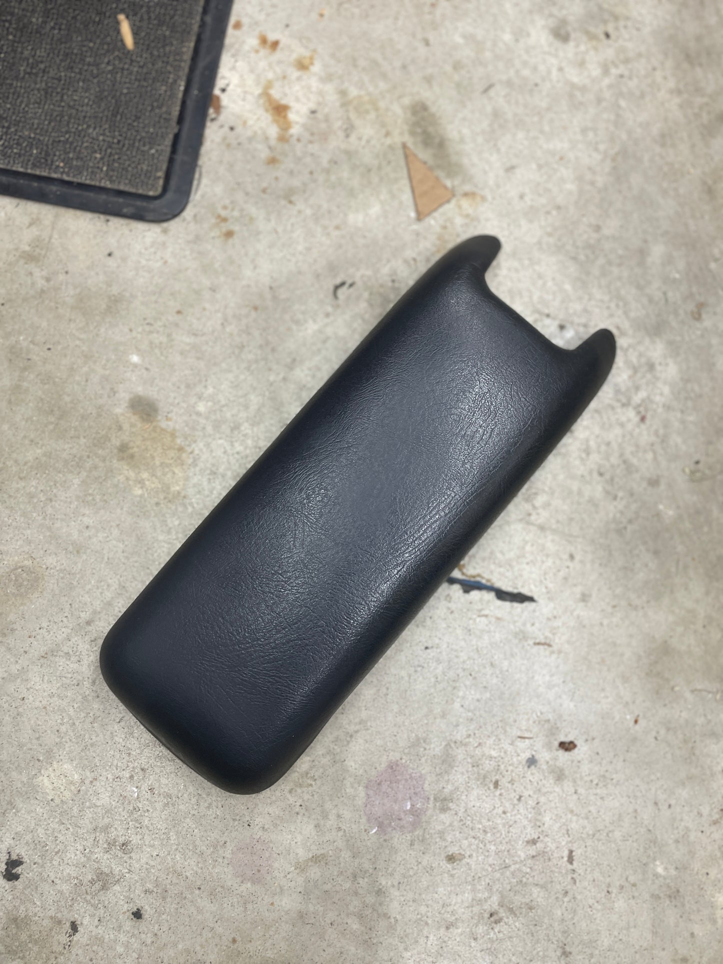 A General Motors OEM Dark Gray Center Console Lid Armrest for 1994-1997 Chevy S10 and Blazer rests on a stained concrete floor, with part of a black mat and small debris nearby.