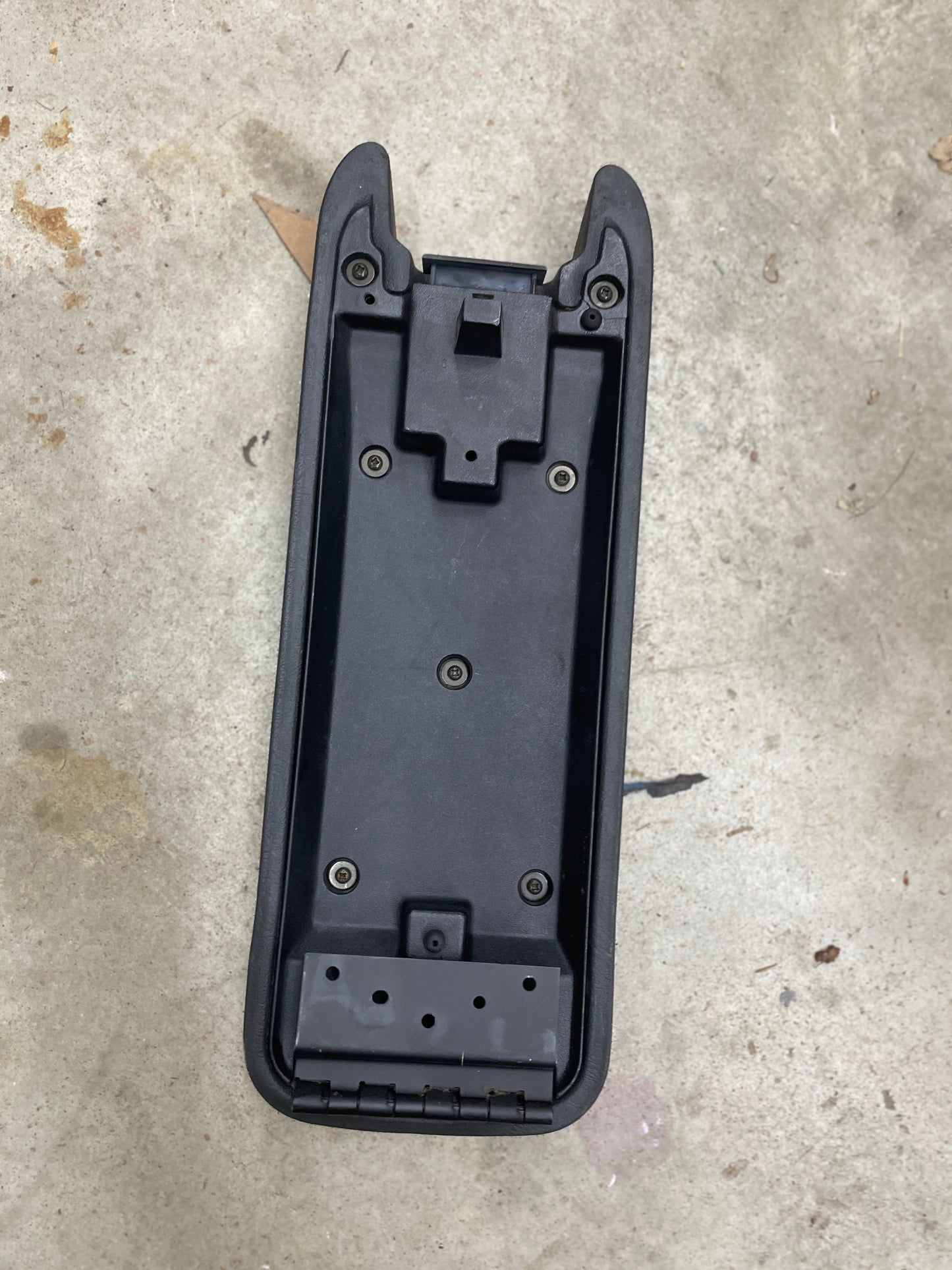 The General Motors OEM Center Console Lid Armrest in Dark Gray fits 1994-1997 Chevy S10, Blazer, and more—ideal as a replacement bracket with screw holes, hinged end, and raised edges. Shown on a concrete surface.