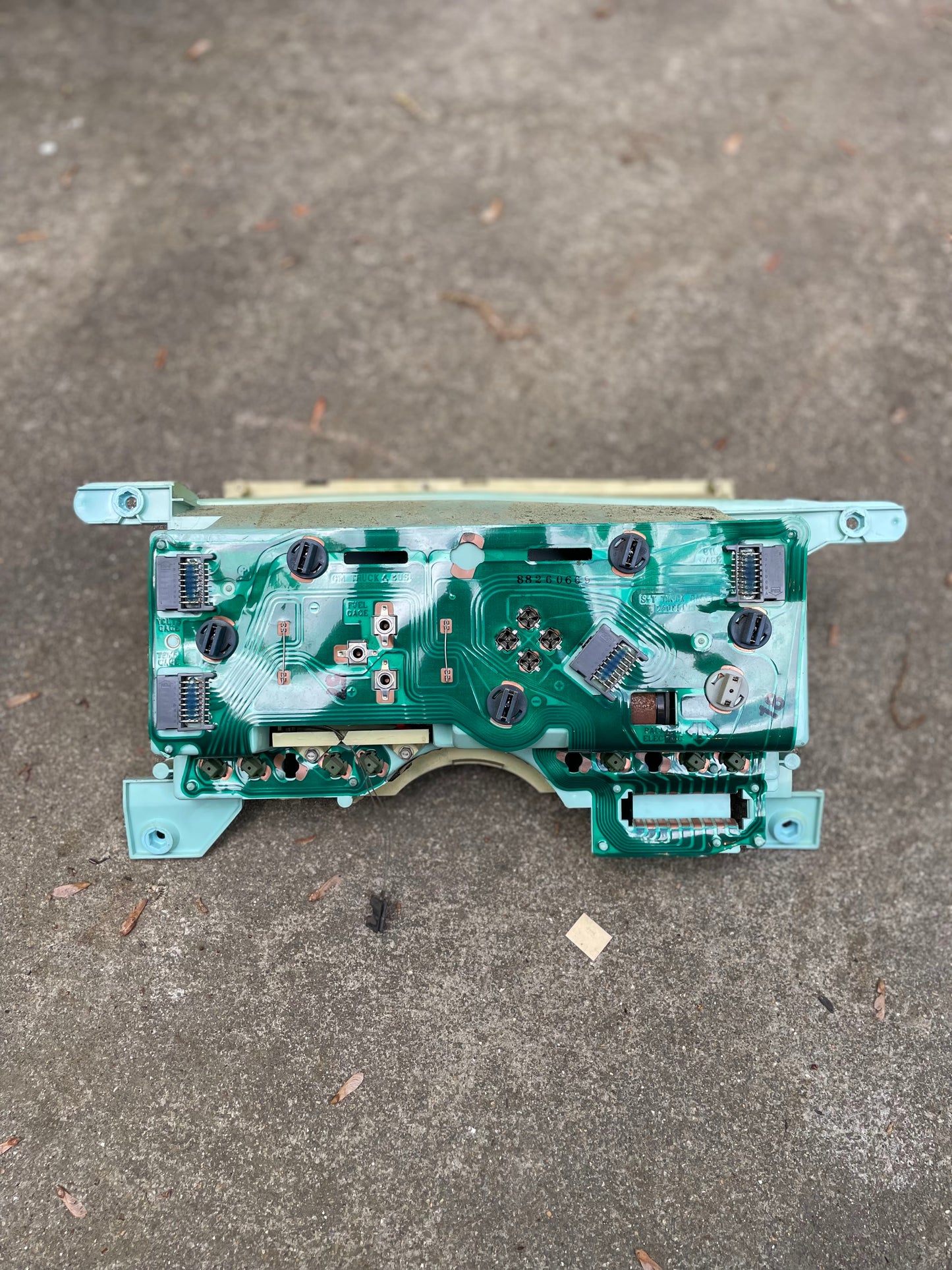 A close-up of a General Motors OEM Instrument Cluster with PRNDL (part #25083728) for 1986-1994 Chevy S10 Blazer and more, resting on rough outdoor concrete with small brown twigs and debris nearby.