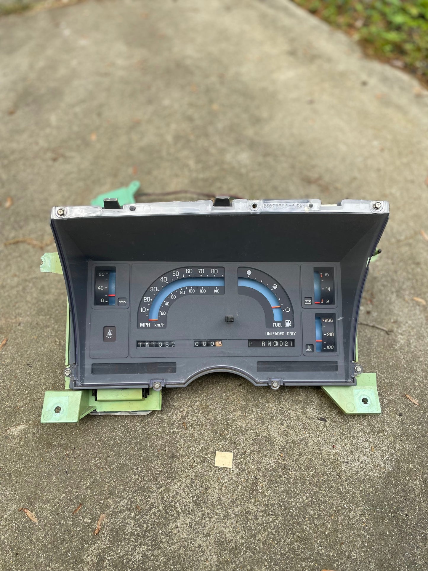A General Motors OEM Instrument Cluster with Mechanical Speedo & PRNDL for 1986-1989 Chevy S10 Blazer is shown resting on a concrete surface outdoors, with grass and leaves visible in the background.