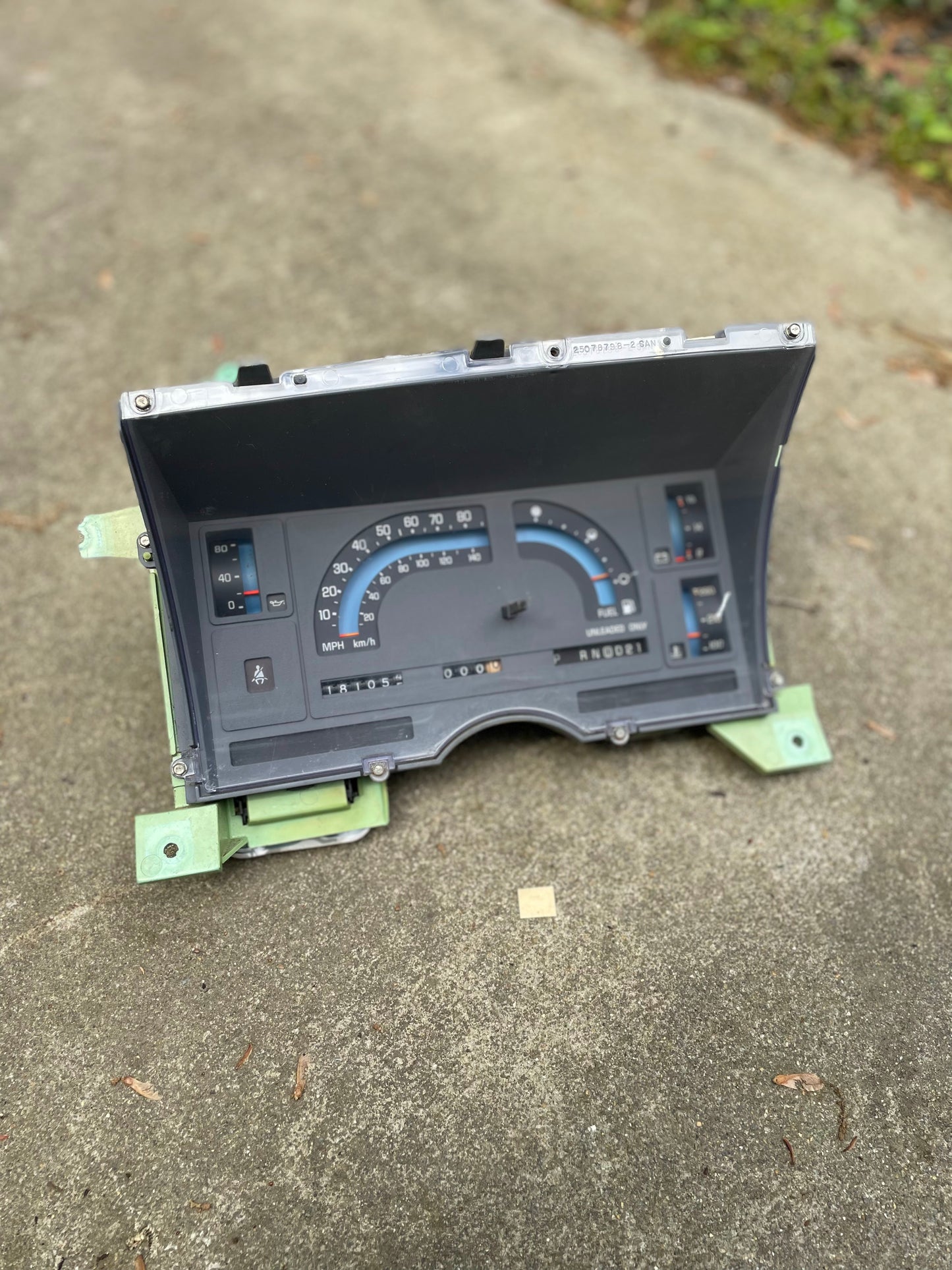 A General Motors OEM Instrument Cluster with mechanical speedo and PRNDL for 1986-1989 Chevy S10 Blazer sits on concrete; fuel gauge and indicator lights are visible, with grass in the background at the top right.