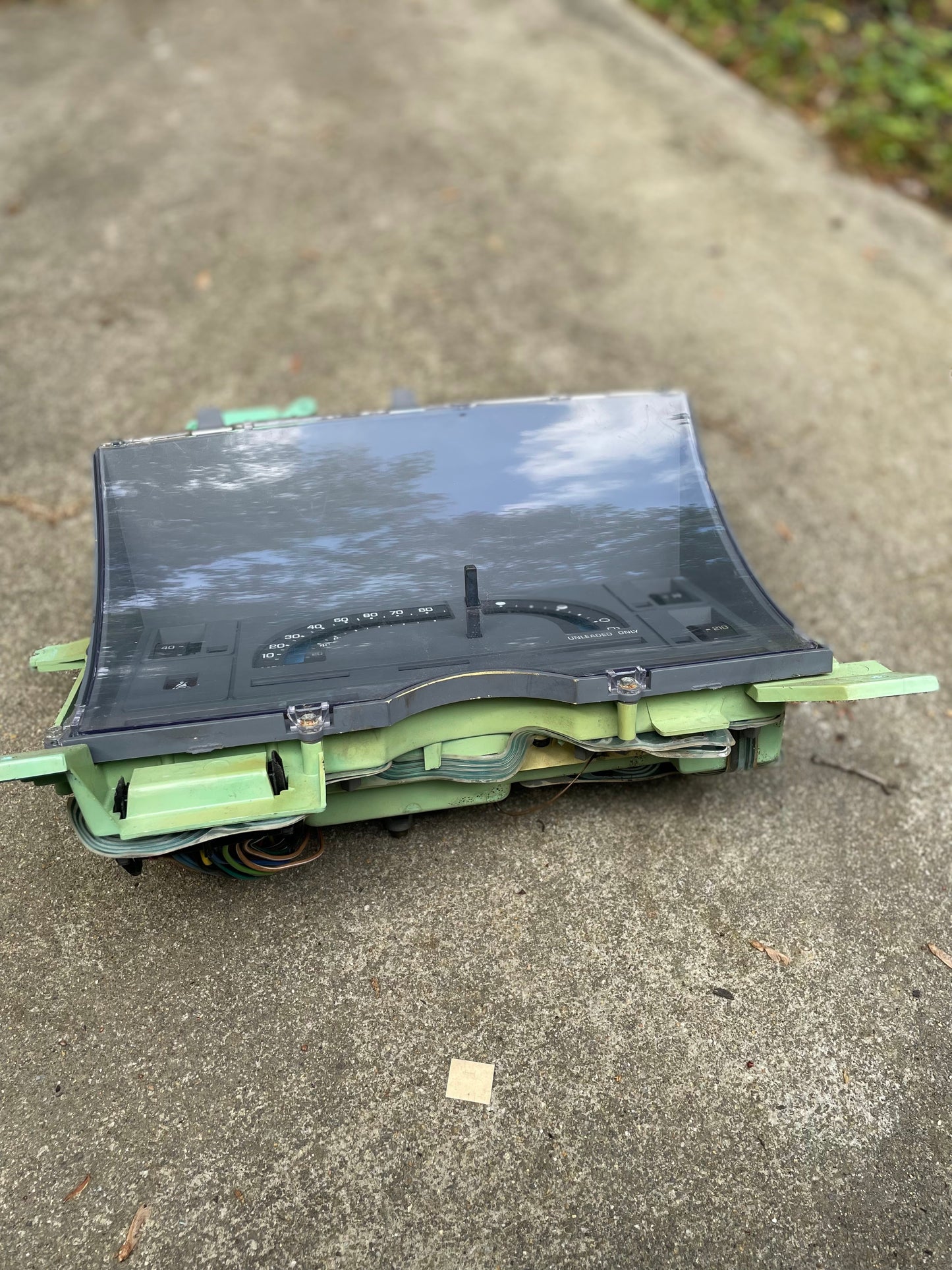 A General Motors OEM Instrument Cluster with Mechanical Speedo & PRNDL for 1986-1989 Chevy S10 Blazer and more sits outdoors on concrete, its curved glass reflecting trees and sky, with visible wires and connectors underneath.