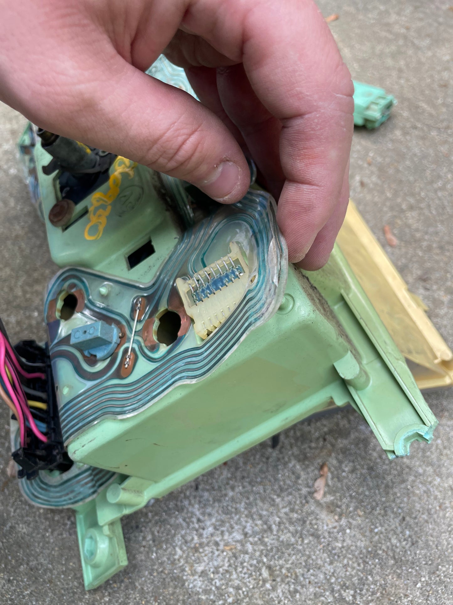 A hand lifts a flexible green circuit board from an old device, revealing a small connector like those in a General Motors OEM Instrument Cluster with Mechanical Speedo & PRNDL for 1986-1989 Chevy S10 Blazer. Background: rough outdoor concrete.