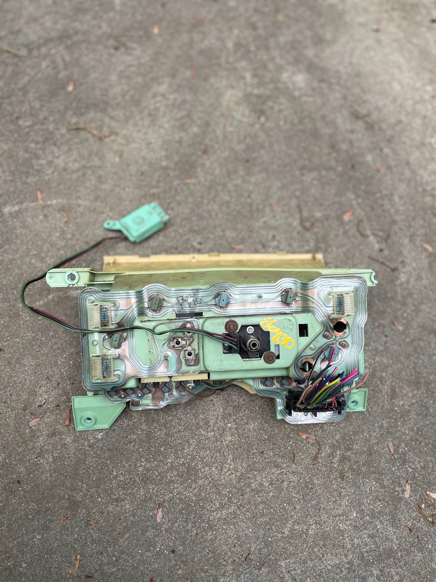 A General Motors OEM Instrument Cluster with Mechanical Speedo & PRNDL for 1986-1989 Chevy S10 Blazer sits on concrete, showing exposed components, wiring, connectors, and a detached plastic piece nearby.