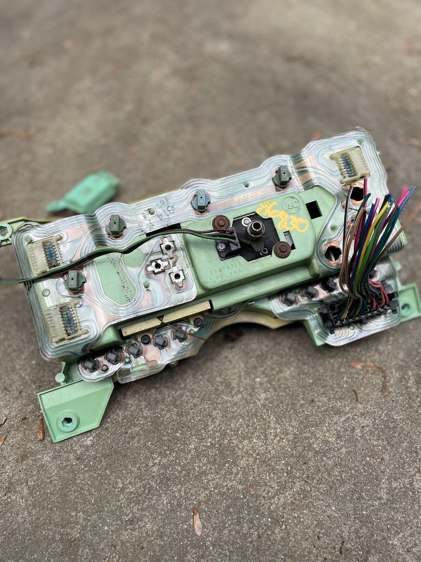 A close-up of a disassembled General Motors OEM Instrument Cluster with Mechanical Speedo & PRNDL for 1986-1989 Chevy S10 Blazer, showing exposed wiring, green and metallic components, connectors, and multicolored wires on concrete.
