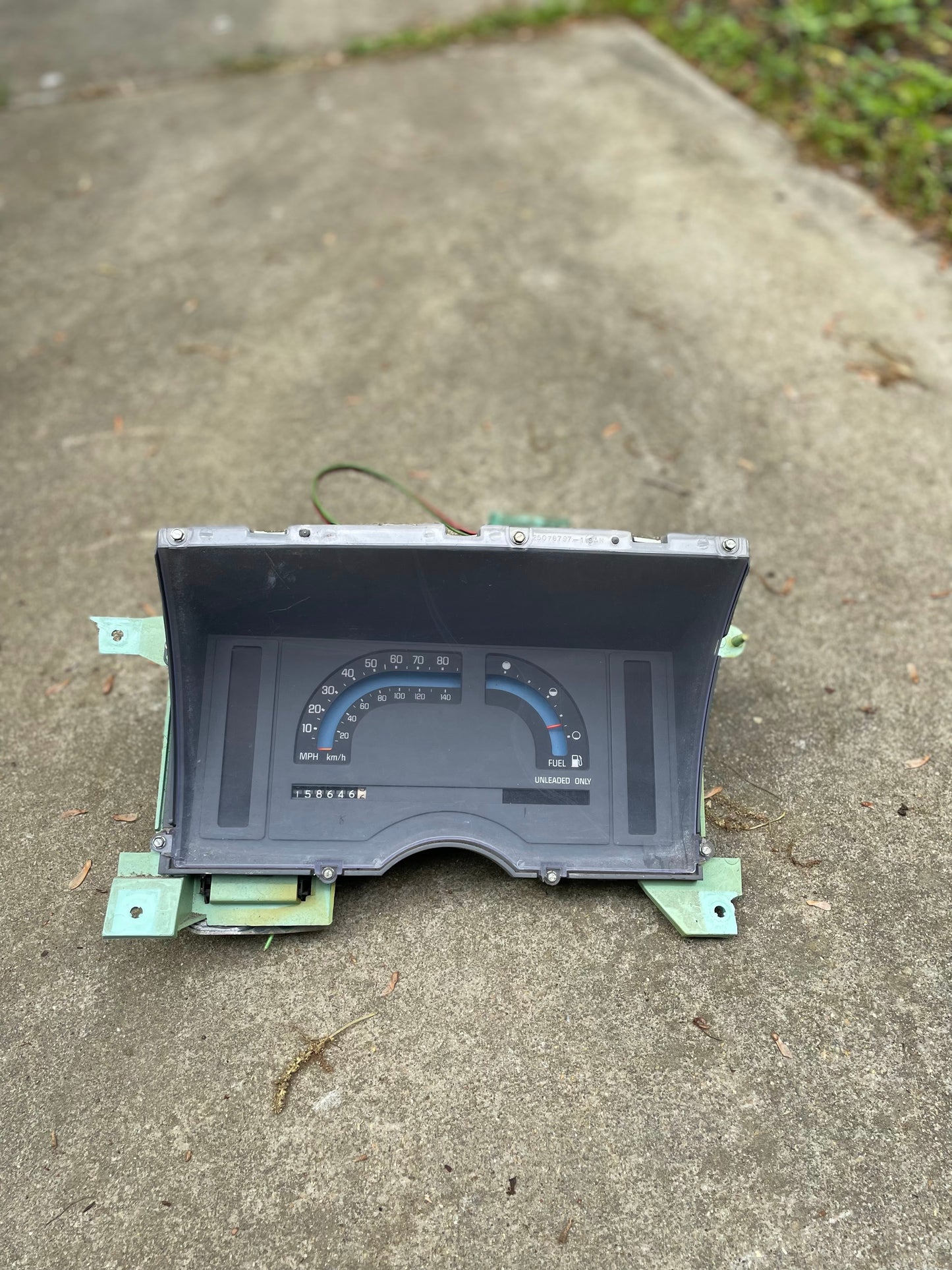 A disassembled General Motors OEM Instrument Cluster with Mechanical Speedo for 1986-1990 Chevy S10 Blazer and manual transmission is shown outdoors on concrete, with green grass visible in the background.