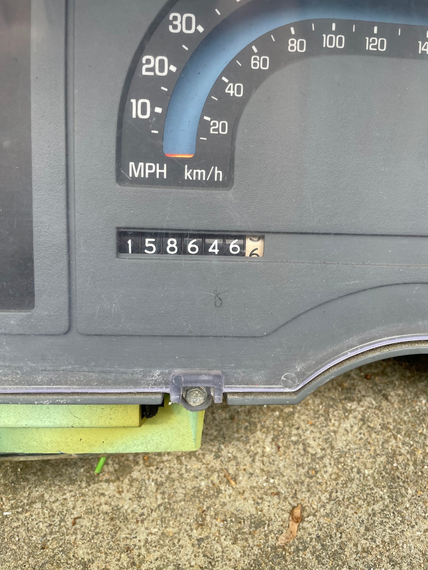 A close-up of a General Motors OEM Instrument Cluster with Mechanical Speedo and Manual Transmission for 1986-1990 Chevy S10 Blazer displays an odometer at 158646 miles, showing both MPH and km/h as the unit rests on concrete.