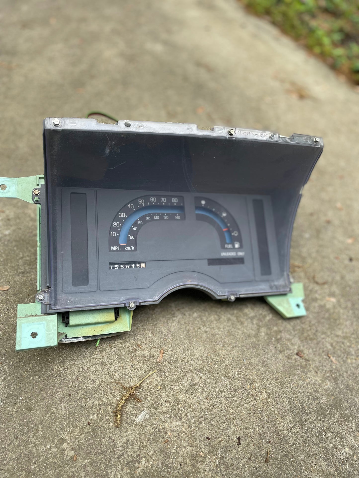 A close-up of a General Motors OEM Instrument Cluster with Mechanical Speedo for 1986-1990 Chevy S10 Blazer, showing both km/h and MPH markings, green metal edges, and manual transmission indicator on concrete.