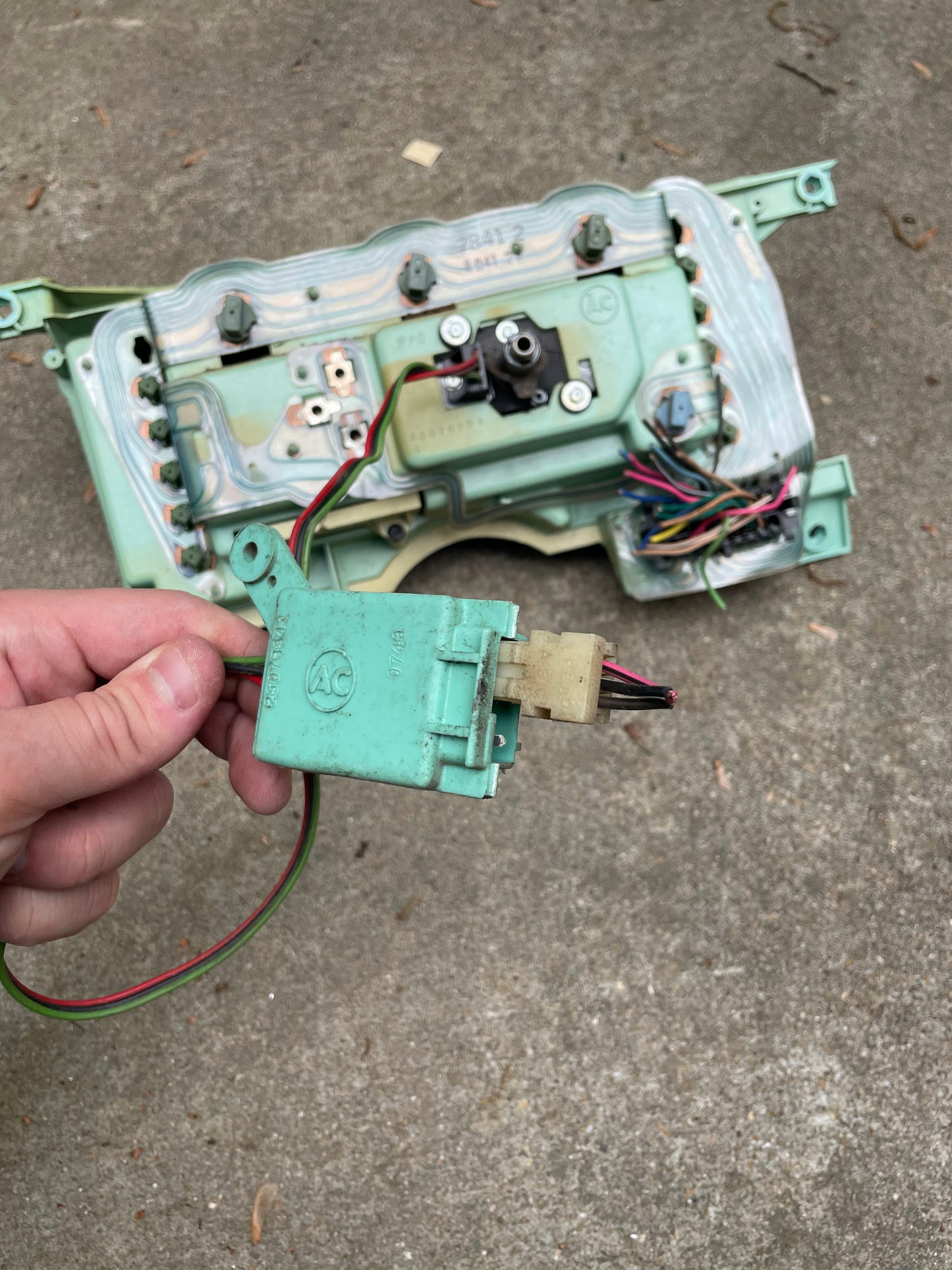 A hand holds a green electrical component with wires in front of a disassembled General Motors OEM Instrument Cluster with Mechanical Speedo for 1986-1990 Chevy S10 Blazer (manual transmission), all laid out on concrete.