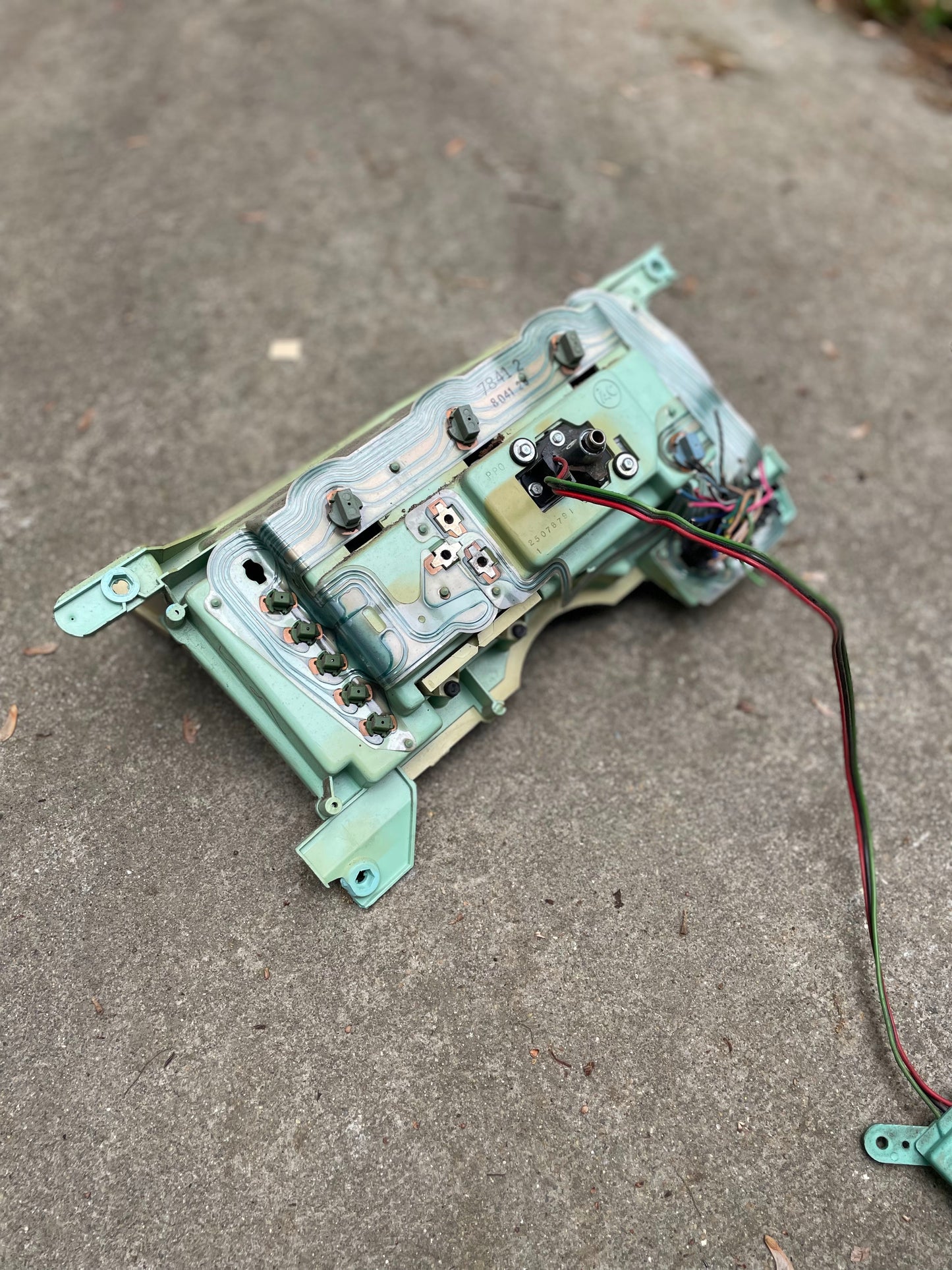 A General Motors OEM Instrument Cluster with Mechanical Speedo for 1986-1990 Chevy S10 Blazer and manual transmission is shown outdoors on concrete, displaying visible wiring and terminals against a softly blurred background.