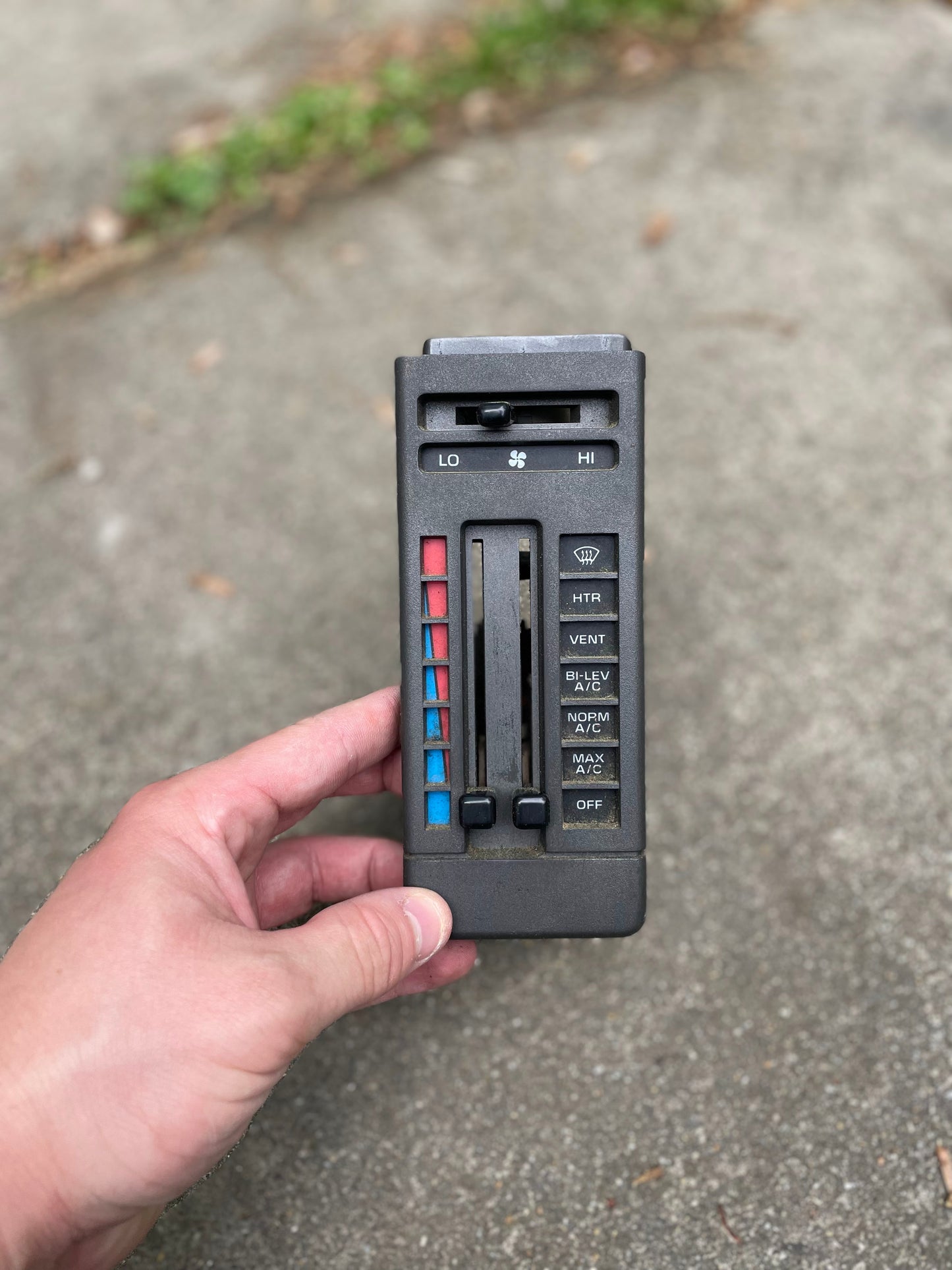 A hand holds a General Motors HVAC Heater Control Panel with A/C for 1986–1994 Chevrolet S10, Blazer and more, featuring sliders and buttons for temperature, fan speed, airflow, and defrost. The background is concrete with grass visible at the top.