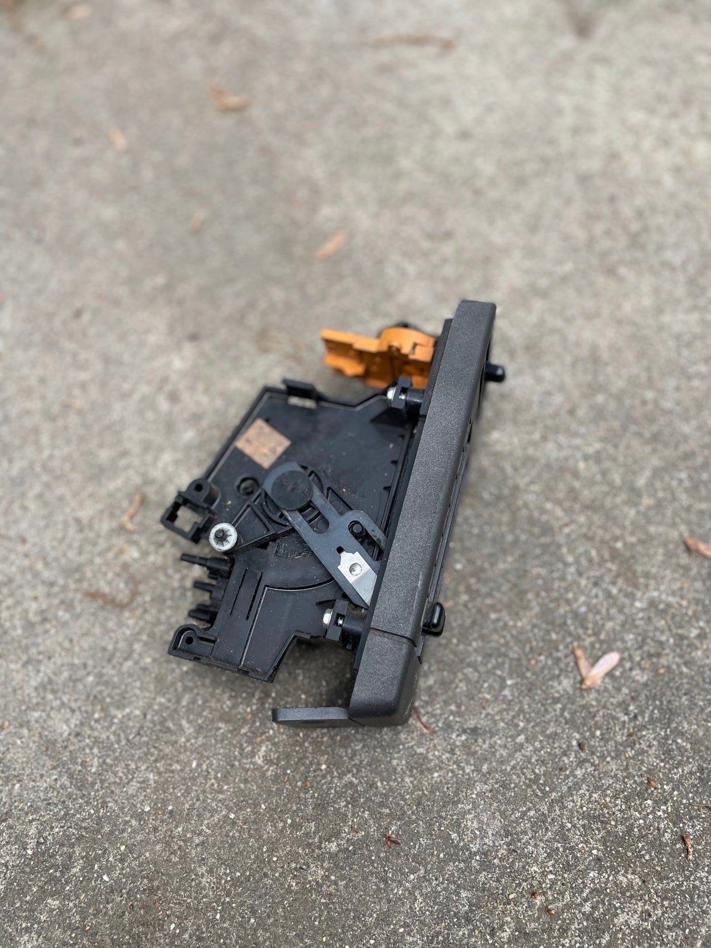 A General Motors HVAC Heater Control Panel with A/C for 1986–1994 Chevrolet S10 and Blazer rests on rough gray concrete, with scattered brown leaves nearby. Its black plastic with metal parts and an orange clip.