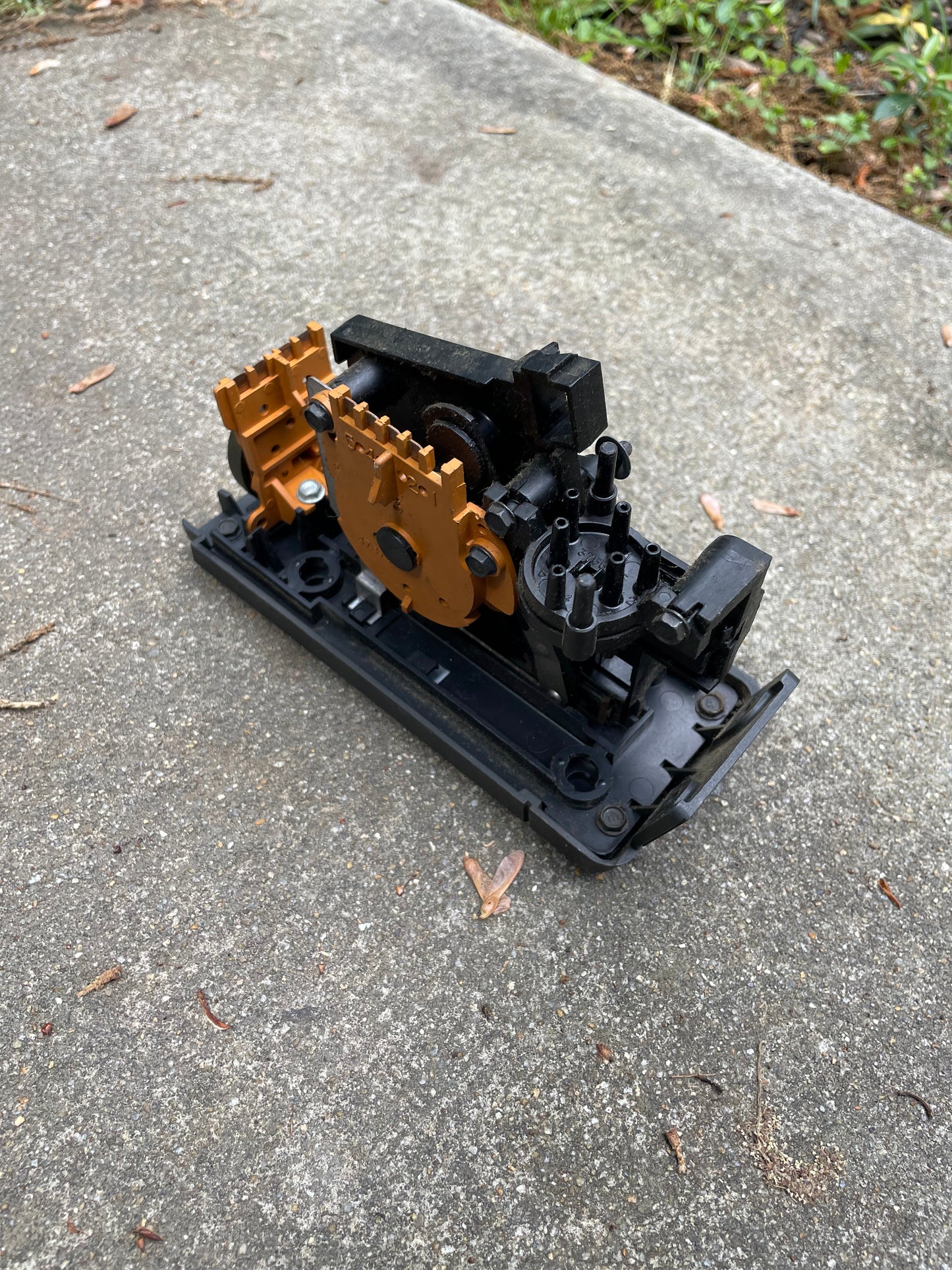 A close-up of a General Motors HVAC Heater Control Panel with A/C for 1986-1994 Chevrolet S10, Blazer, and more, showing its black and orange plastic gears and levers on an outdoor concrete surface with grass in the background.