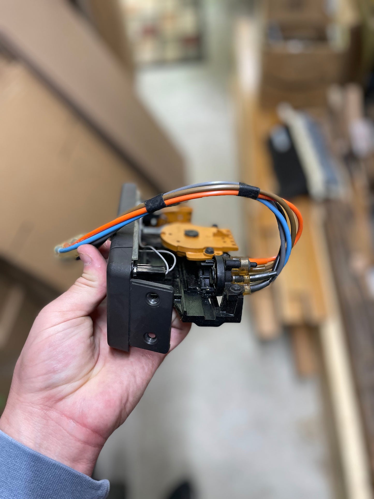 A hand holds a General Motors HVAC Heater Control Panel with A/C for 1986-1994 Chevrolet S10 and Blazer, with orange, blue, and silver wires attached—set before shelves and wooden boards in the background.