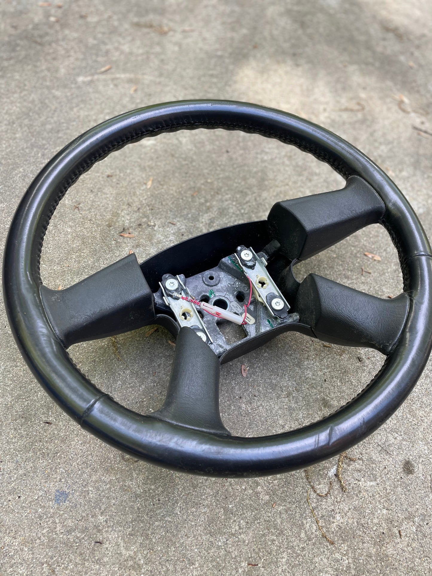 An OEM Leather Steering Wheel by General Motors for 2002-2009 GM Chevy Silverado, Trailblazer, and more is shown outdoors on a rough concrete surface, with exposed metal and wires visible in the center.