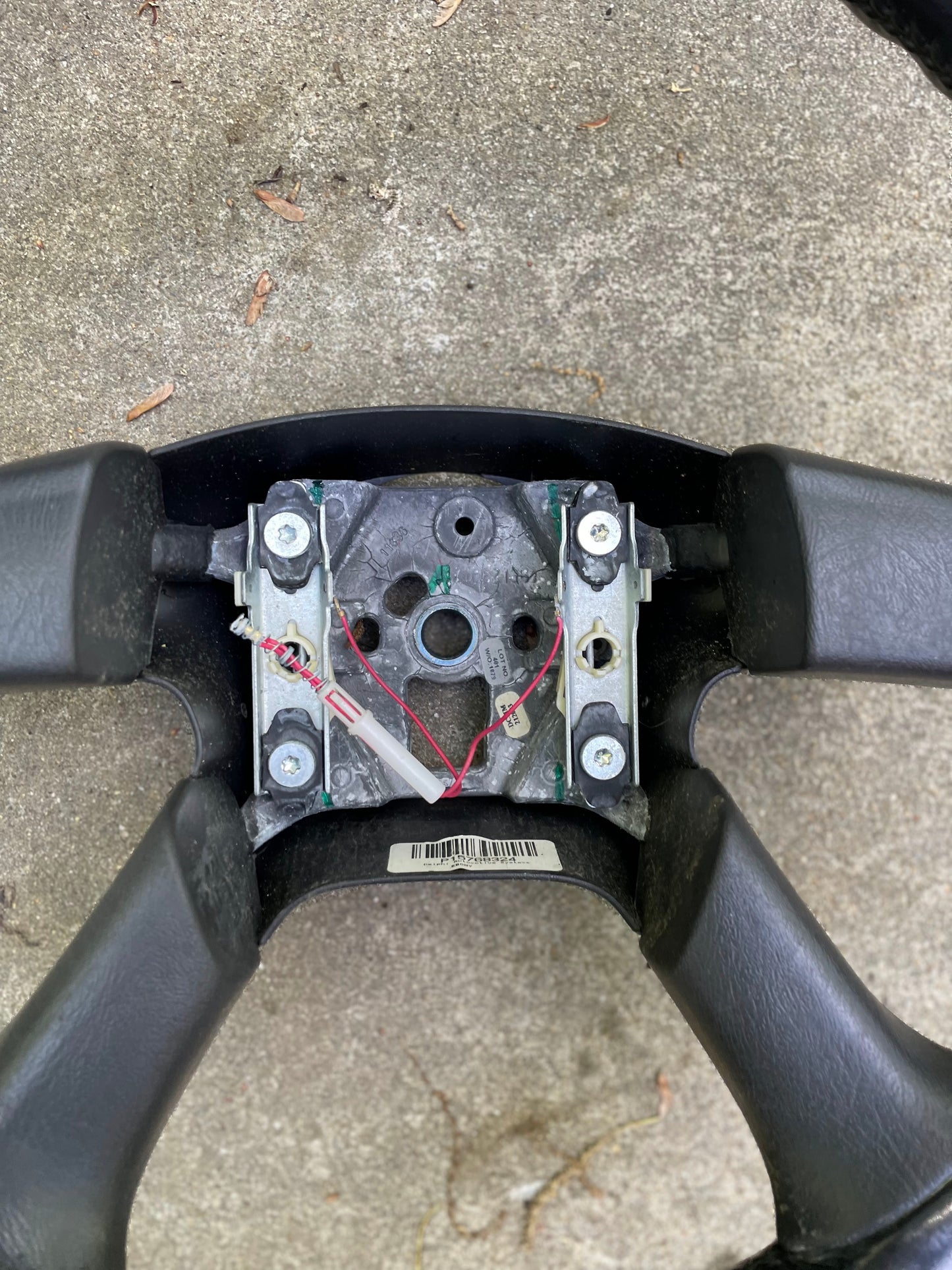A close-up of a General Motors OEM Leather Steering Wheel for 2002-2009 Chevy Silverado, Trailblazer, and more, shown with the center cover removed to reveal metal brackets, screws, and colorful wires against a concrete background.