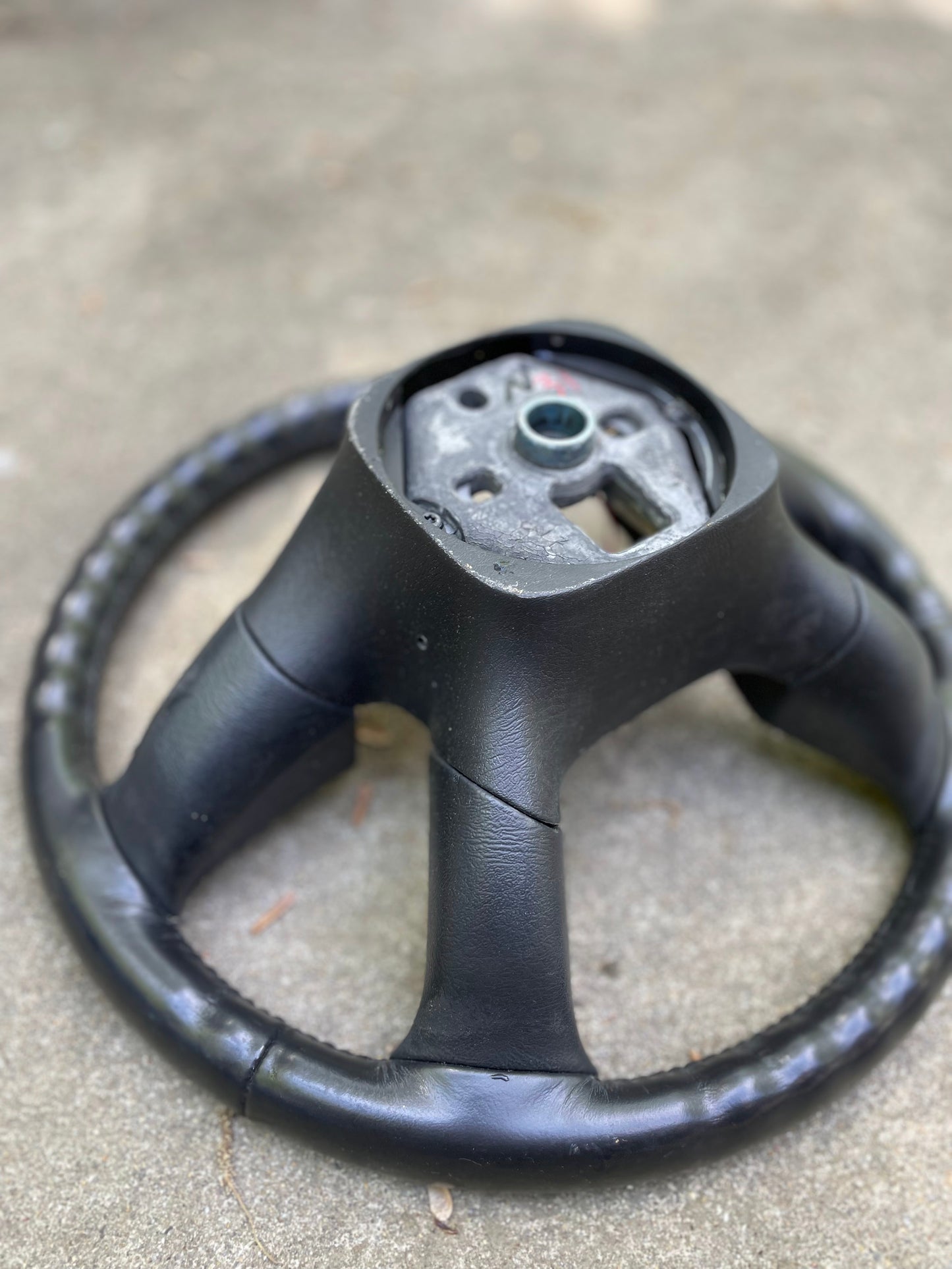 A General Motors OEM Leather Steering Wheel for 2002-2009 Chevy Silverado, Trailblazer, and more lies detached on a light-colored concrete surface, showing its exposed center hub and mounting components.