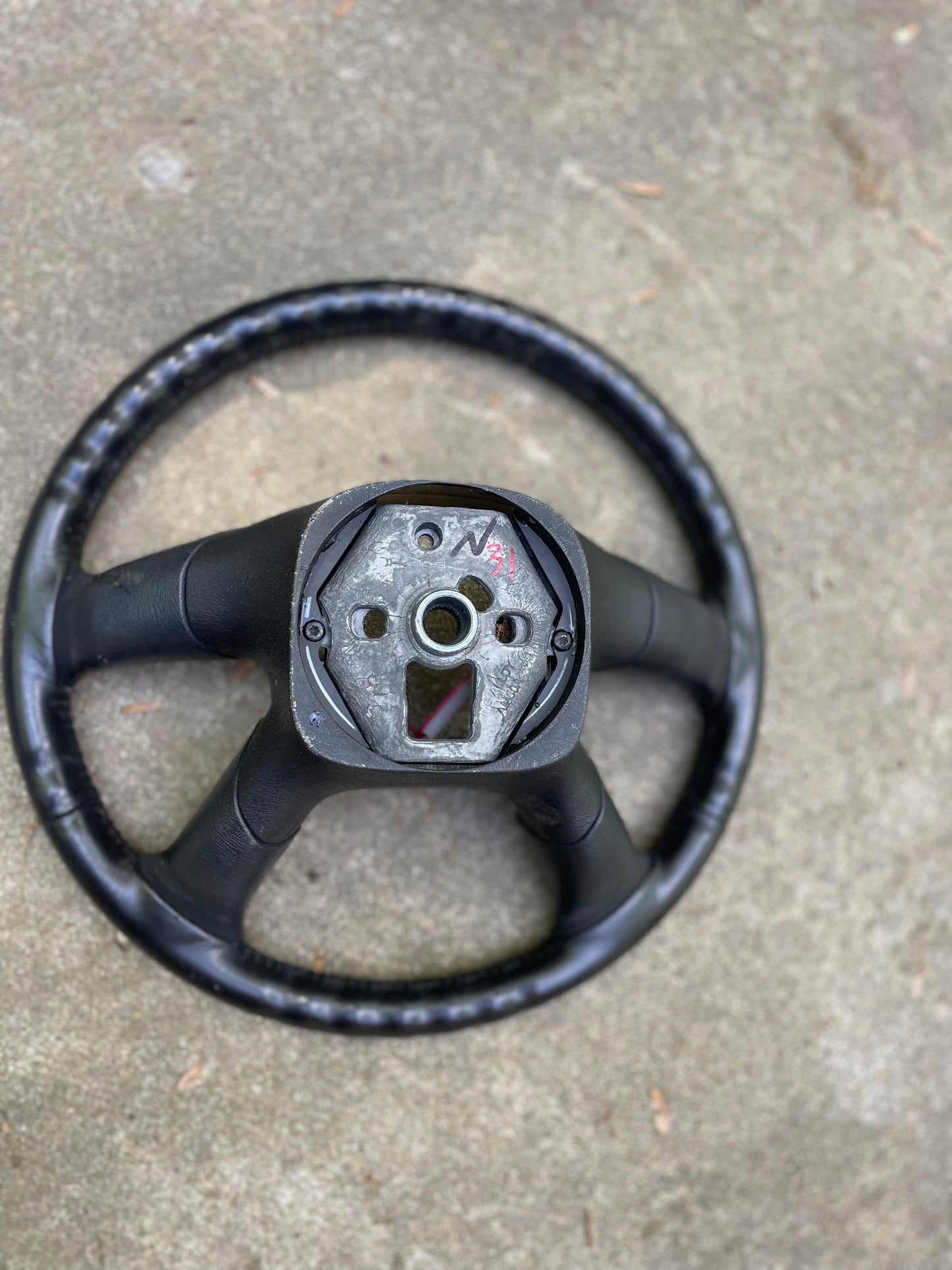 A used OEM Leather Steering Wheel by General Motors for 2002-2009 GM Chevy Silverado, Trailblazer, and more rests on rough concrete, showing visible metal parts and screw holes with worn black covering.
