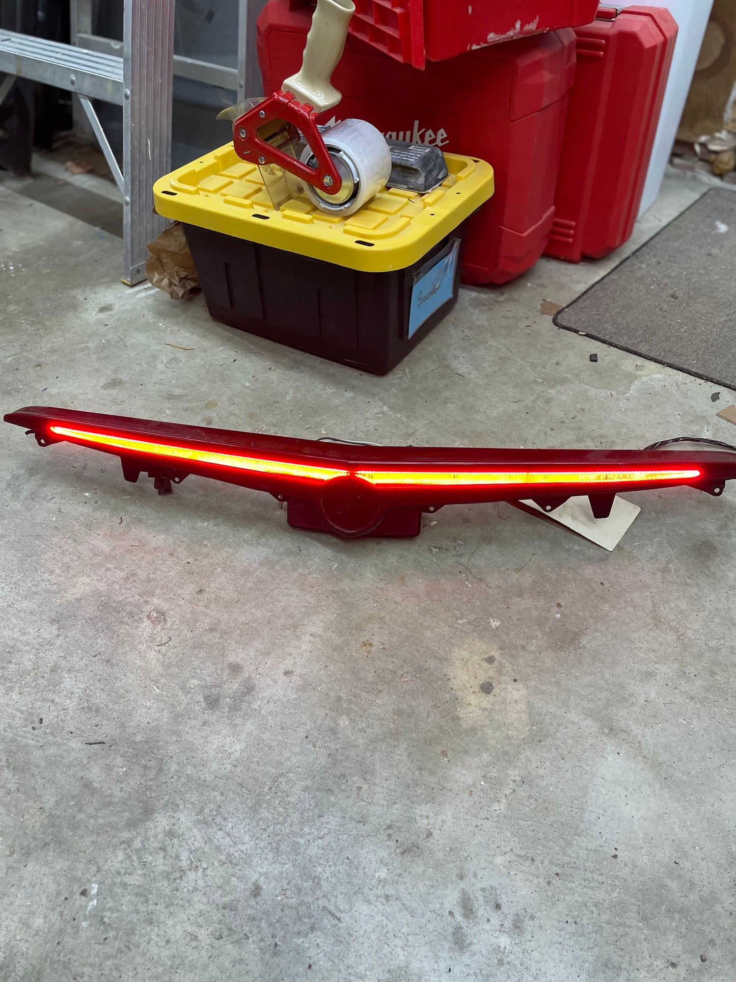 A General Motors OEM Center High Mount Third Brake Lamp CHMSL for 2003-2007 Cadillac CTS lies illuminated on a concrete garage floor, with storage bins, tape, and tools nearby—ready for replacing your vehicle’s third brake light.