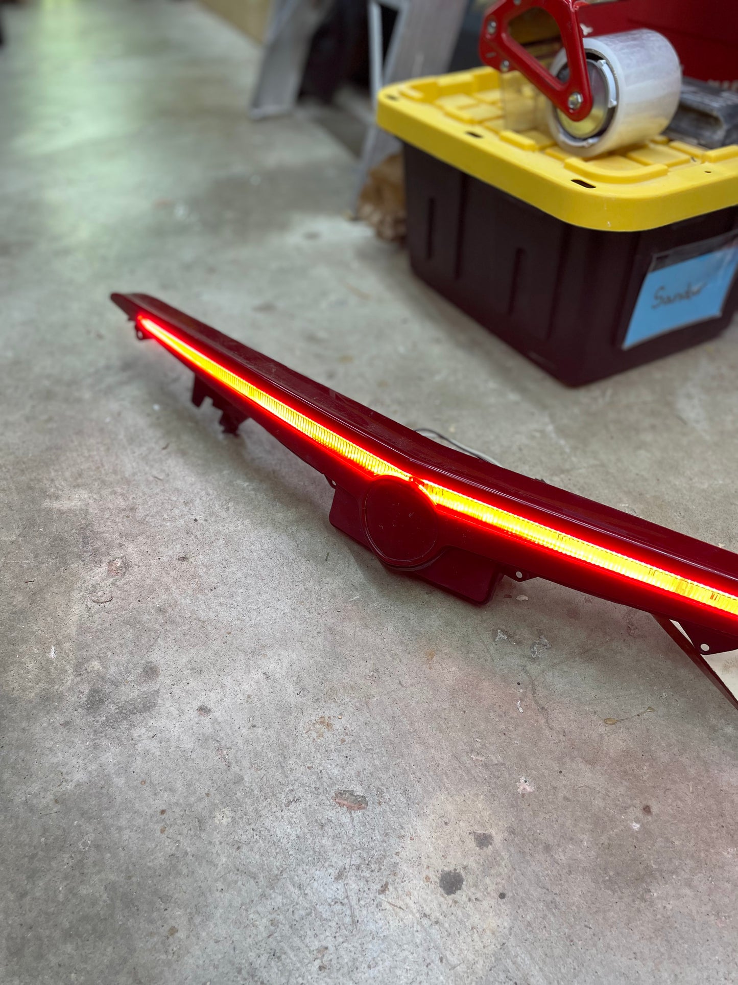 An OEM Center High Mount Third Brake Lamp CHMSL for 2003-2007 Cadillac CTS by General Motors glows red on a garage floor, with a black and yellow storage bin and packing tape dispenser in the background.