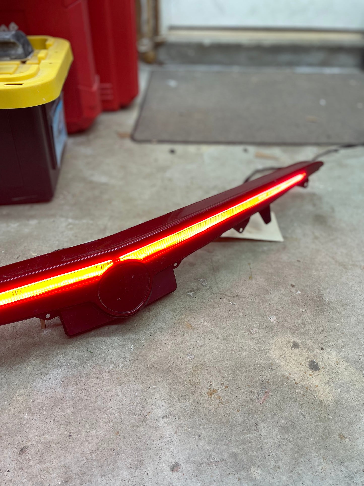 An illuminated OEM Center High Mount Third Brake Lamp (CHMSL) for 2003-2007 Cadillac CTS by General Motors, featuring a yellow LED strip, rests on a concrete garage floor near a black and yellow storage bin.