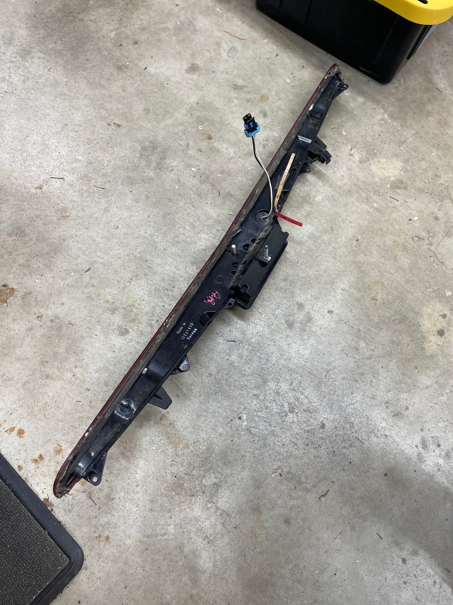 A General Motors OEM Center High Mount Third Brake Lamp (CHMSL) for 2003-2007 Cadillac CTS with exposed wires lies on a concrete garage floor, with part of a yellow storage bin visible in the corner.
