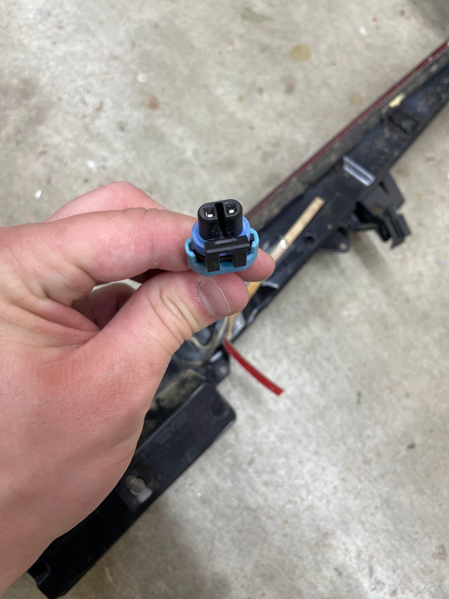 A hand holds a black and blue electrical connector near an OEM Center High Mount Third Brake Lamp (CHMSL) for 2003-2007 Cadillac CTS by General Motors, with a metal and plastic assembly visible against a concrete floor background.