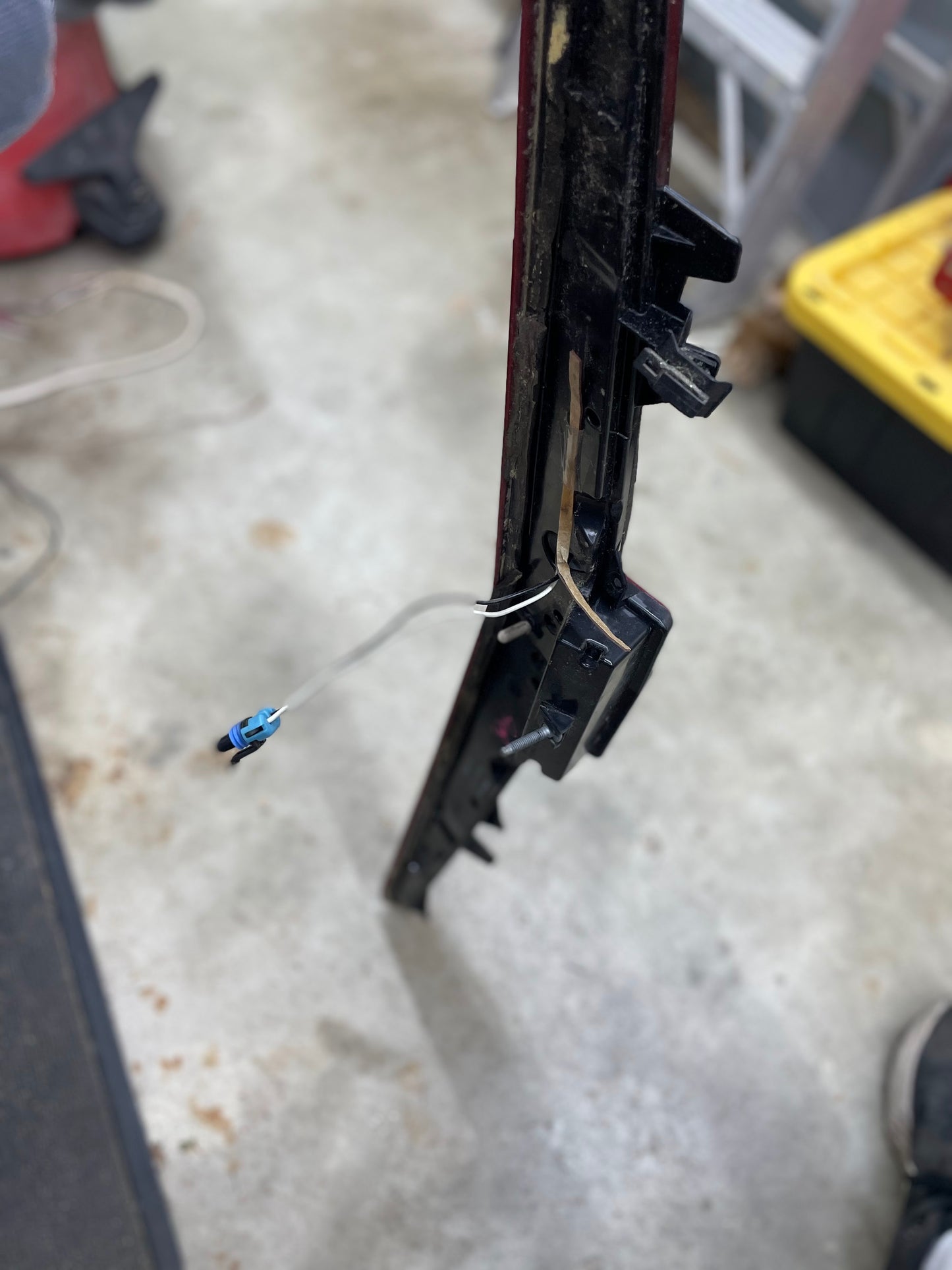 Close-up of a General Motors OEM Center High Mount Third Brake Lamp (CHMSL) for 2003-2007 Cadillac CTS with exposed wire and blue connector, resting on a garage floor. Tools and storage bins are blurred in the background.