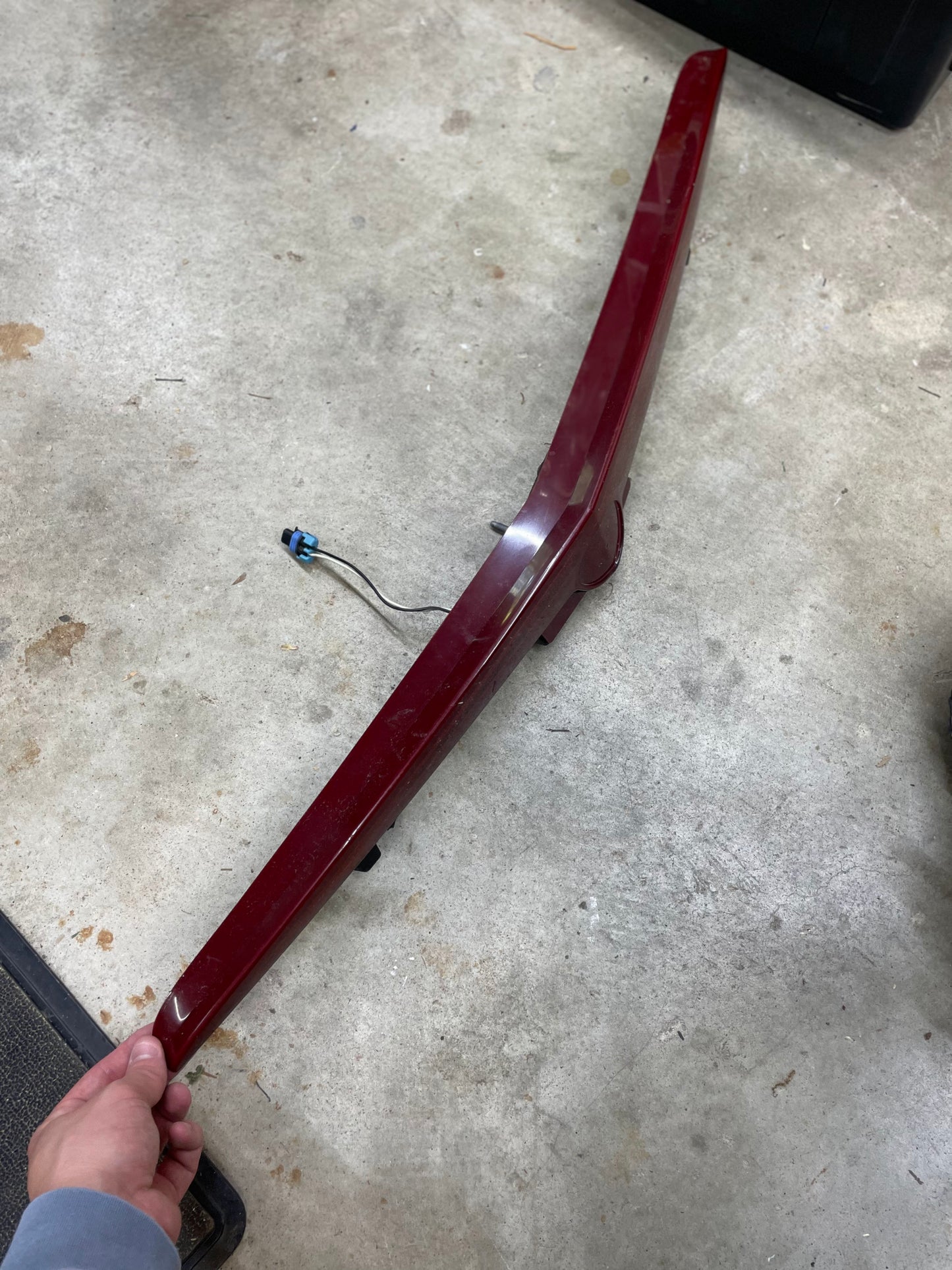 A person holds a long, red General Motors OEM Center High Mount Third Brake Lamp (CHMSL) for 2003-2007 Cadillac CTS with an attached electrical connector, resting on a concrete floor.