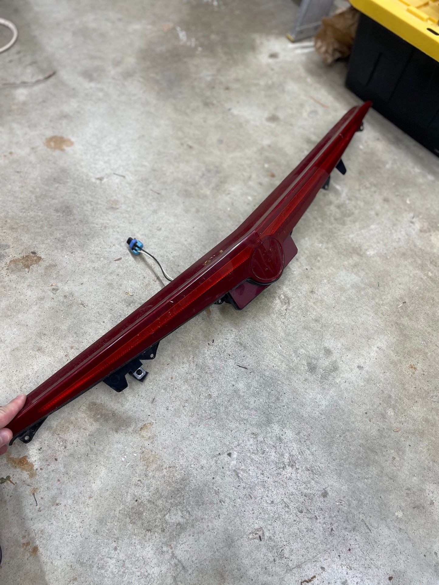 A person holds an OEM Center High Mount Third Brake Lamp CHMSL for 2003-2007 Cadillac CTS by General Motors, with a yellow and black storage bin and tools visible in the background on a concrete floor.