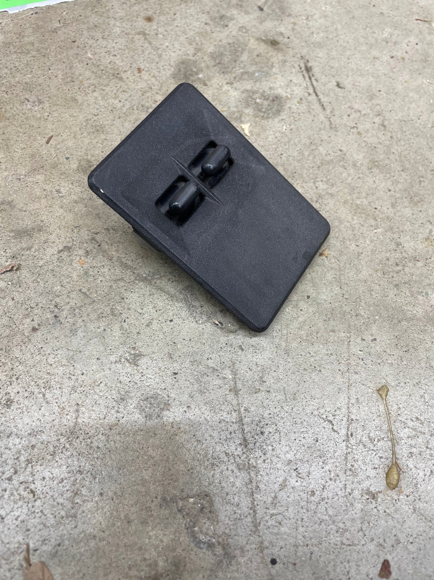 A black, rectangular plastic cover with two top clips from a General Motors OEM Driver Side Window Switch for 2 Door 1986-94 Chevy S10, Blazer, Syclone, and more lies on rough, light gray concrete.